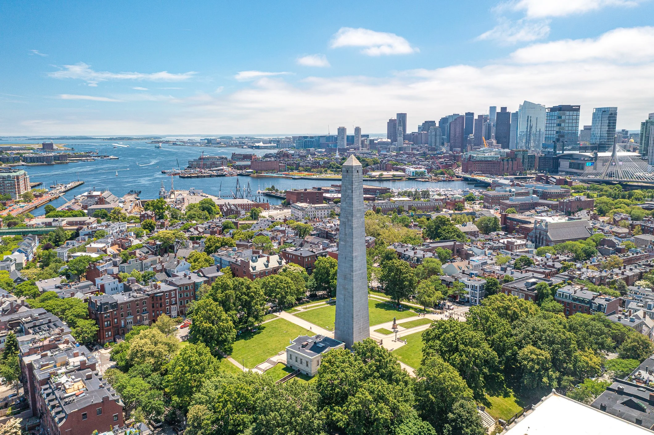 Aerial view of Boston with the Boston Obelisk in the foreground, harbor in the background, and city skyline under a blue sky with clouds.