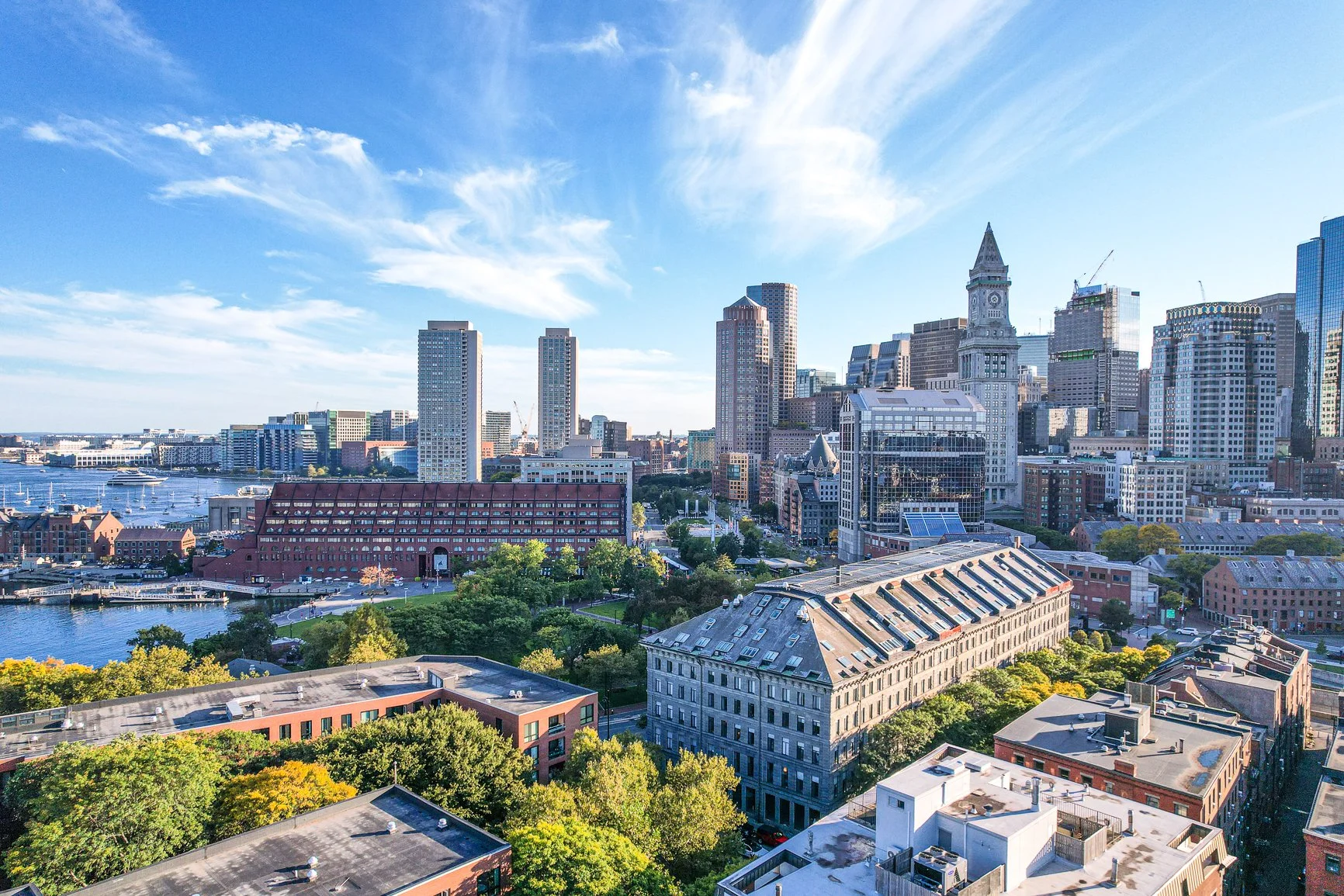 A city skyline featuring tall skyscrapers, historic buildings, a waterfront with boats, and lush green trees in the foreground under a partly cloudy blue sky.