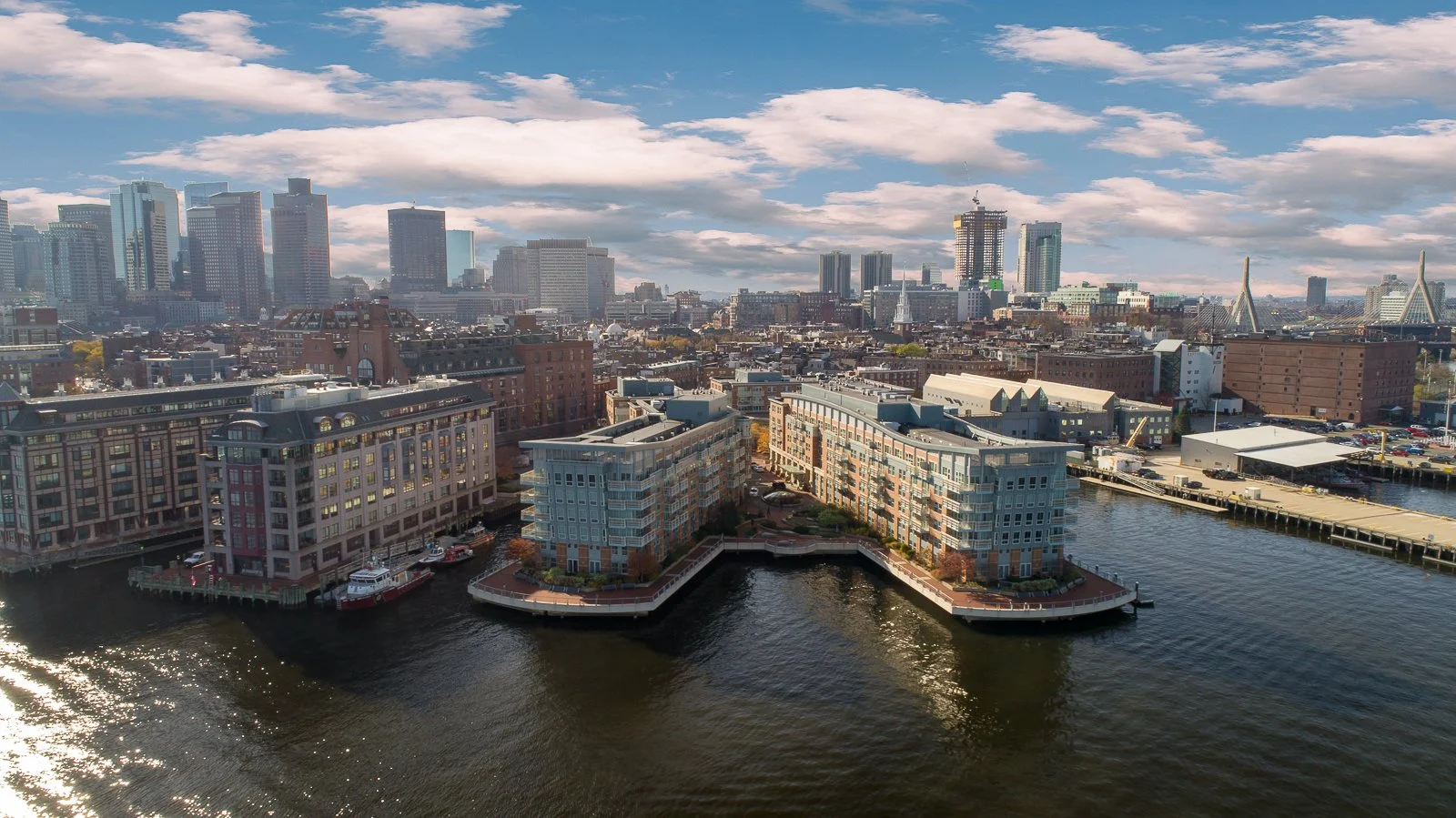 A city skyline with tall modern buildings in the background, and waterfront residential buildings on the water's edge in the foreground, under a partly cloudy sky.