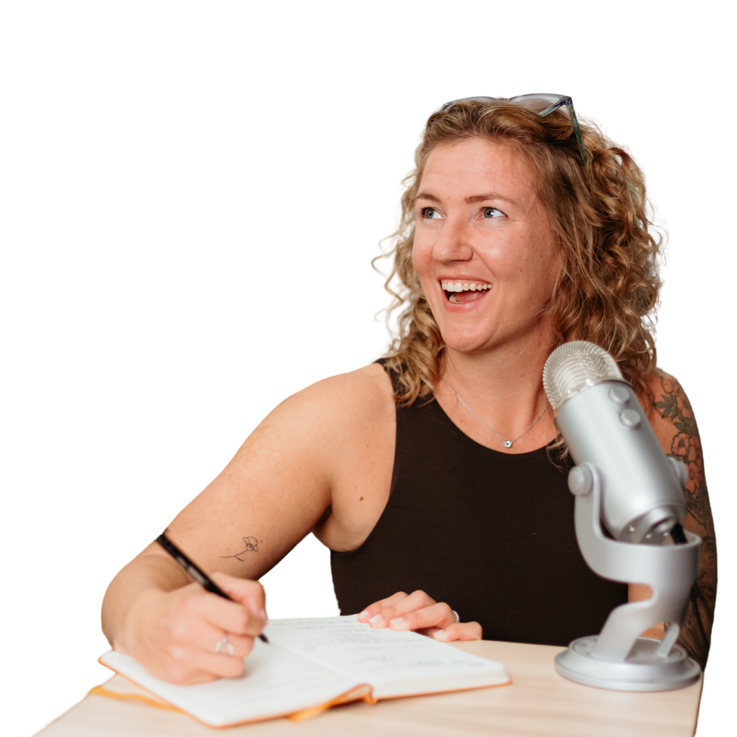 Sam at nomad copy, in a brown tank top, writing in a journal while speaking into a podcasting microphone