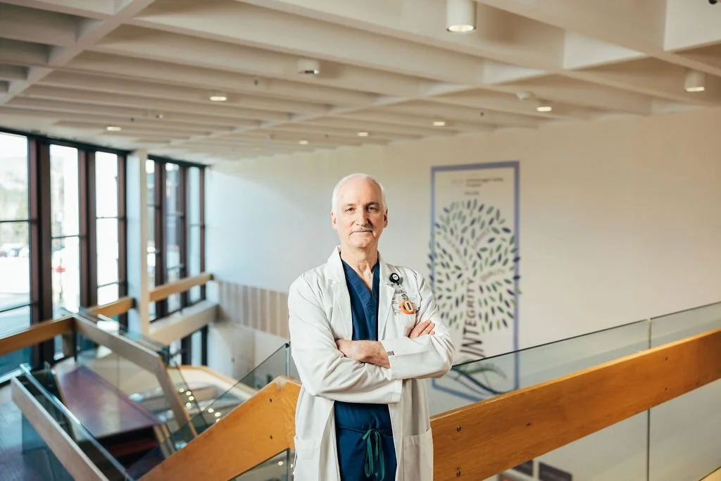 An elderly male doctor standing on a staircase landing inside a hospital or medical facility, wearing a white coat and stethoscope, with arms crossed and looking at the camera.