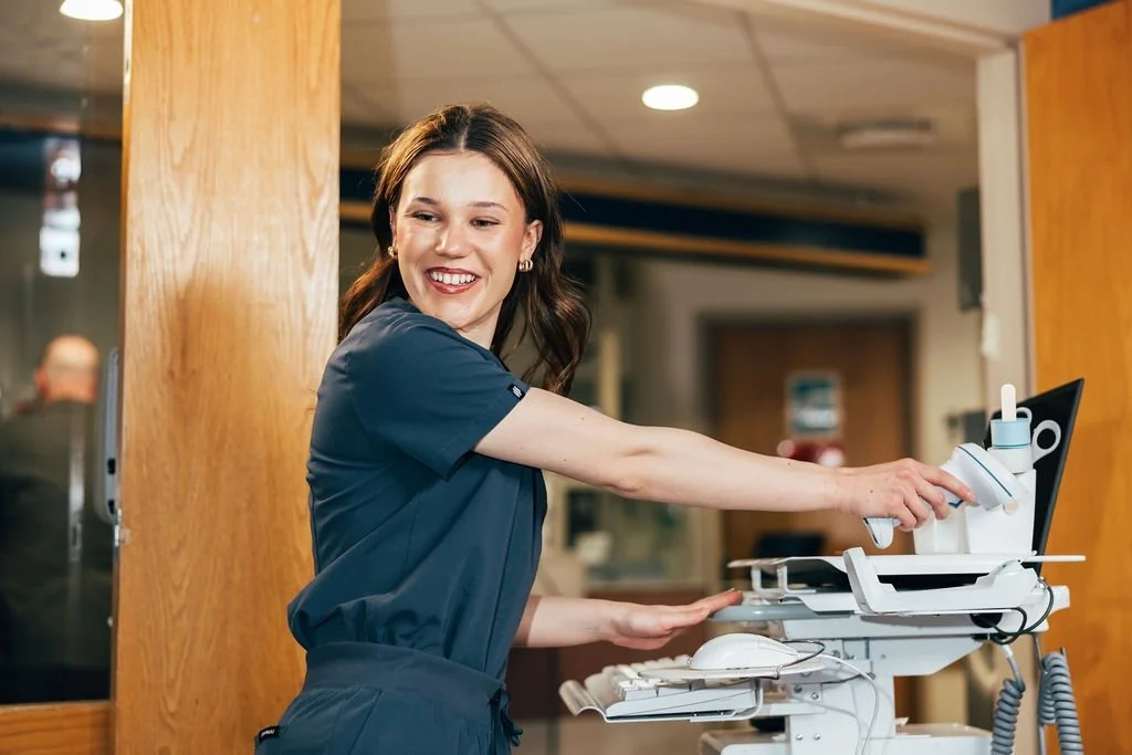A female healthcare worker in navy scrubs smiling and adjusting medical equipment on a cart in a hospital or clinic setting.