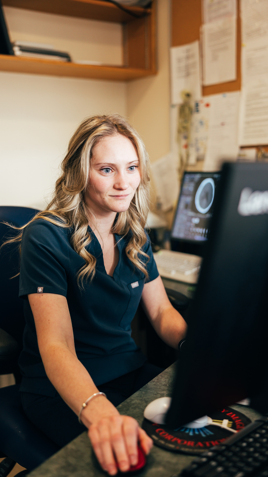 A woman wearing dark scrubs working at a computer in an office setting.