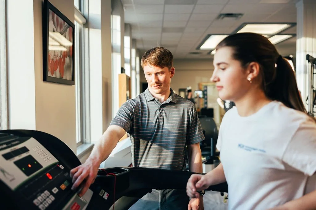 A man and a woman at a gym, with the man on a treadmill and the woman adjusting the settings.