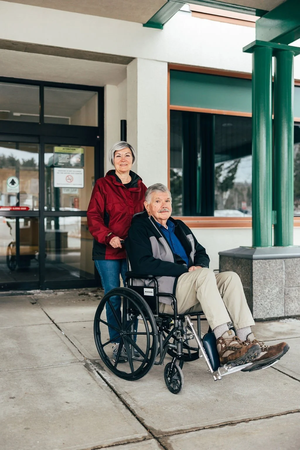 An elderly man in a wheelchair and a woman standing behind him outside a building, possibly a medical or care facility, during daytime.
