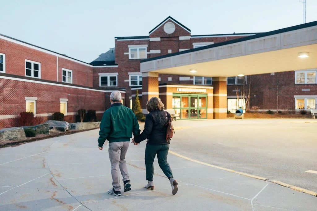 An elderly couple holding hands and walking in front of a hospital entrance at dusk.