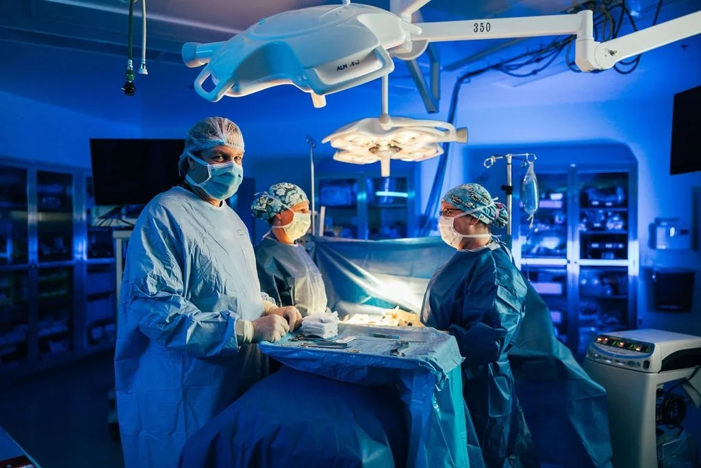 Medical team performing surgery in an operating room with bright surgical lights, medical equipment, and surgeons wearing scrubs, masks, and caps.