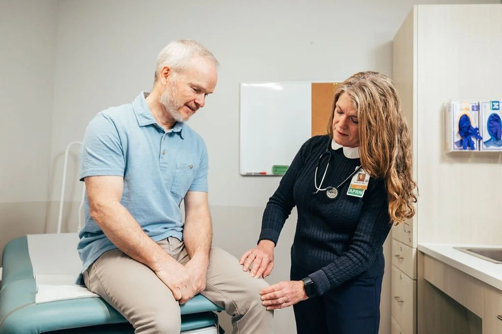 A patient sitting on an examination table in a medical office, talking to a female nurse or doctor who is examining his leg. The healthcare professional has a stethoscope around her neck and is wearing a name badge that says 'APR.  N.