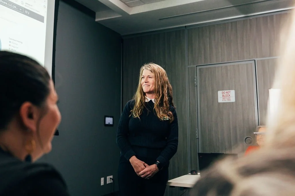 A woman with long brown hair and a black sweater standing in front of a gray wall, smiling during a presentation or meeting in a conference room with other seated attendees.