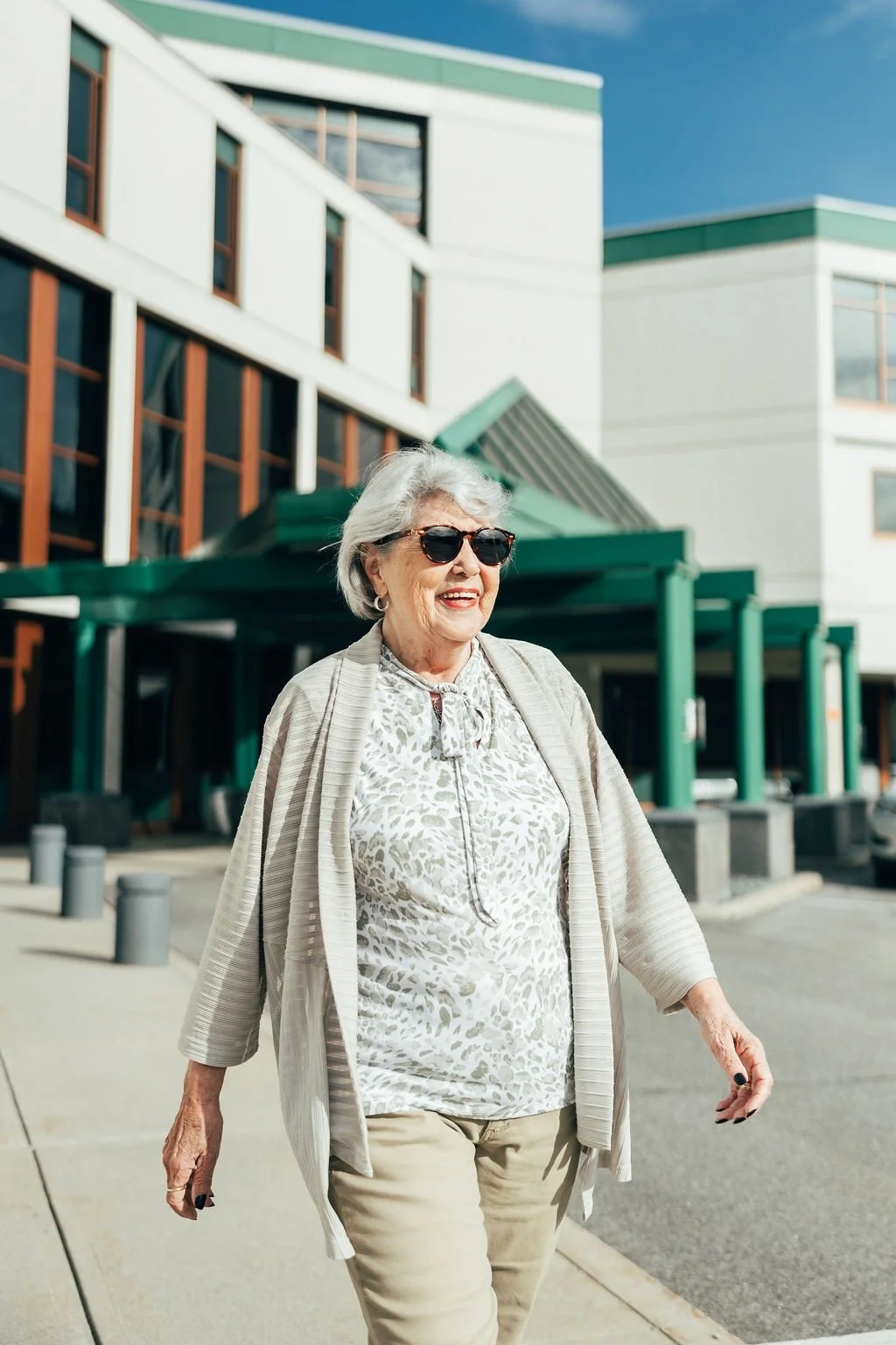 An elderly woman with gray hair, wearing sunglasses, a patterned blouse, beige pants, and a light cardigan, walking outdoors with a modern building in the background.