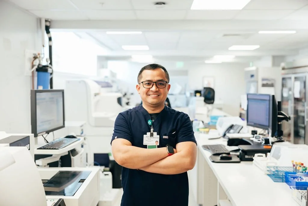 A smiling man in a navy blue scrub top with a hospital ID badge, glasses, and a smartwatch, standing with arms crossed in a busy medical laboratory with computer monitors and lab equipment.
