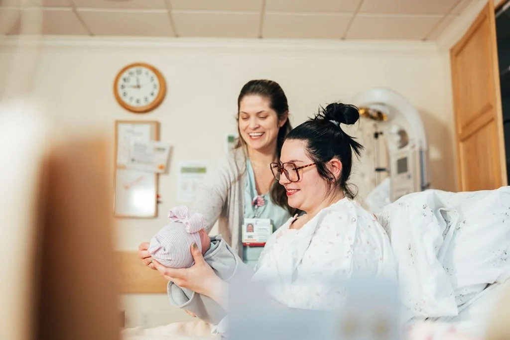 A woman holding a newborn baby in a hospital room, with a nurse or medical professional smiling beside her.