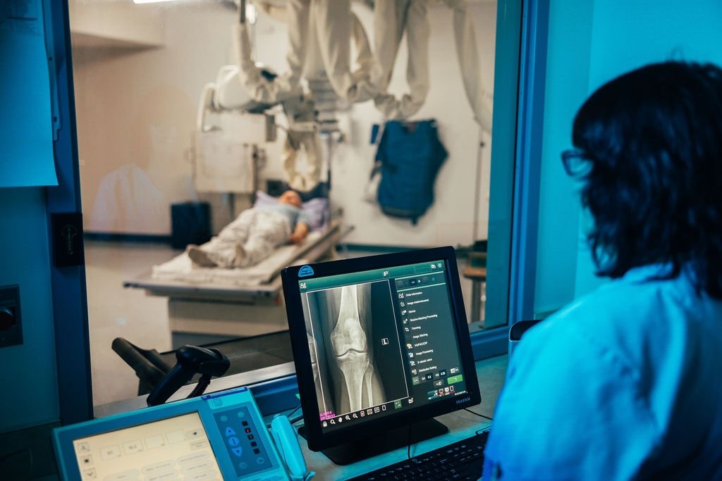 Medical professional examining an X-ray on a computer screen in a hospital's radiology room, with a patient lying on a bed in the background.