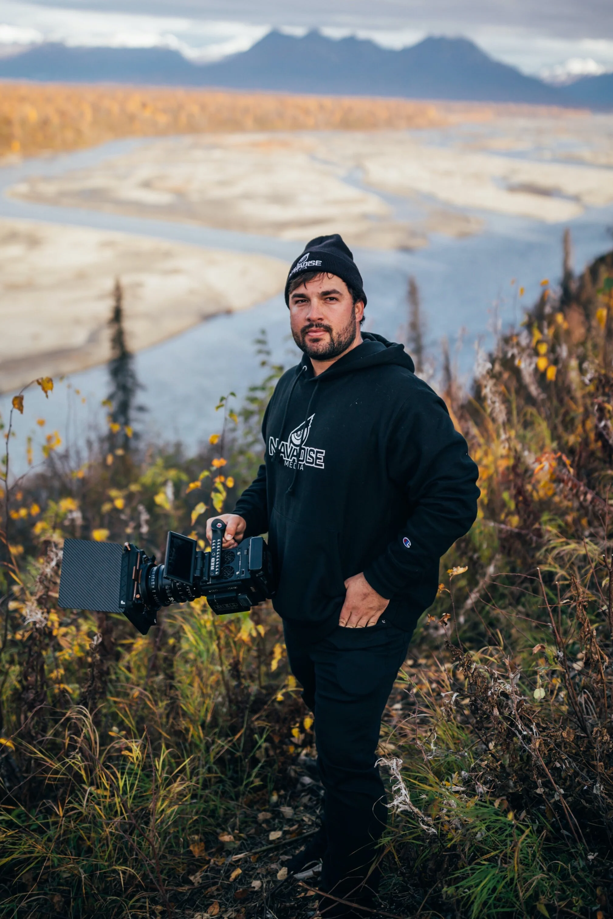 Photo of Casey Nava of Navadise Media, Casey is holding a camera with mountains and stream in background