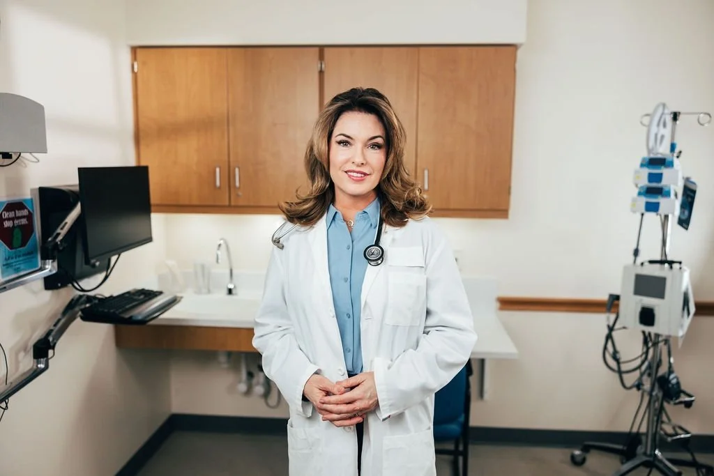 Female doctor in white coat standing in a medical examination room with test equipment and a monitor.