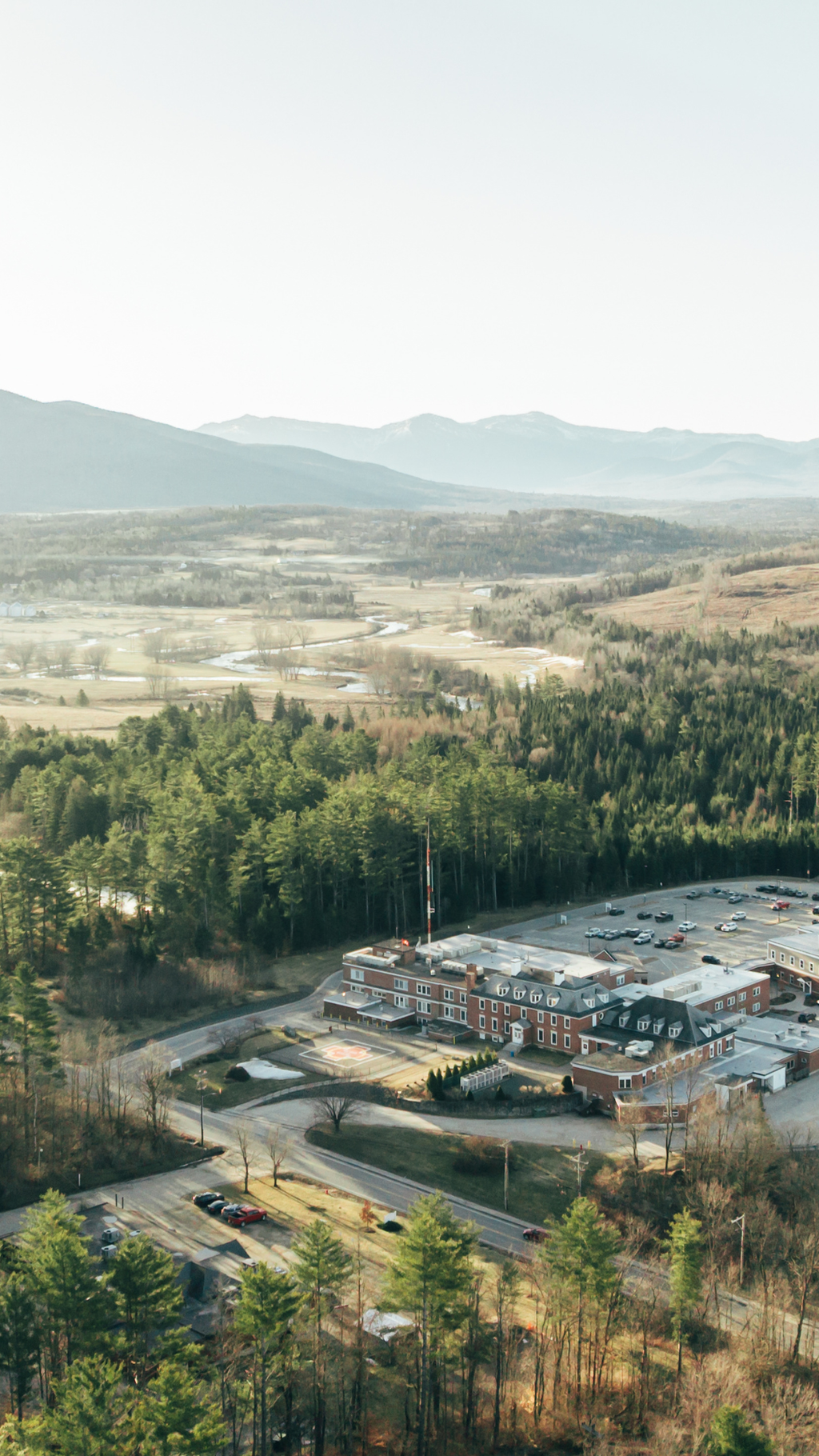 An aerial view of a hospital building surrounded by parking lots, trees, and distant mountains.