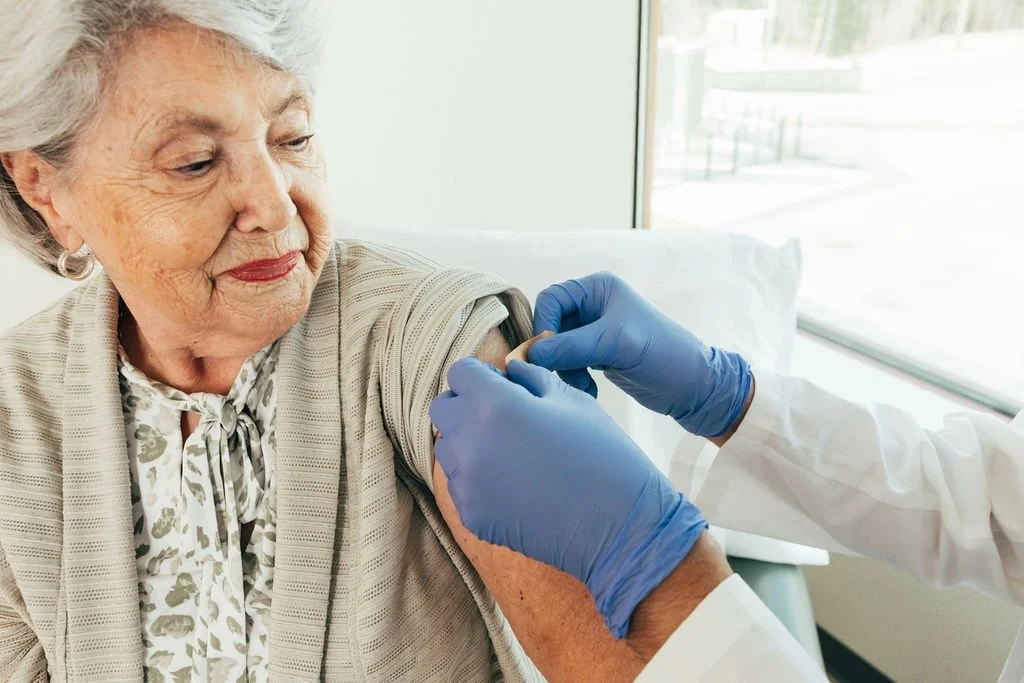 An elderly woman receiving a vaccination in her upper arm from a healthcare professional wearing blue gloves.