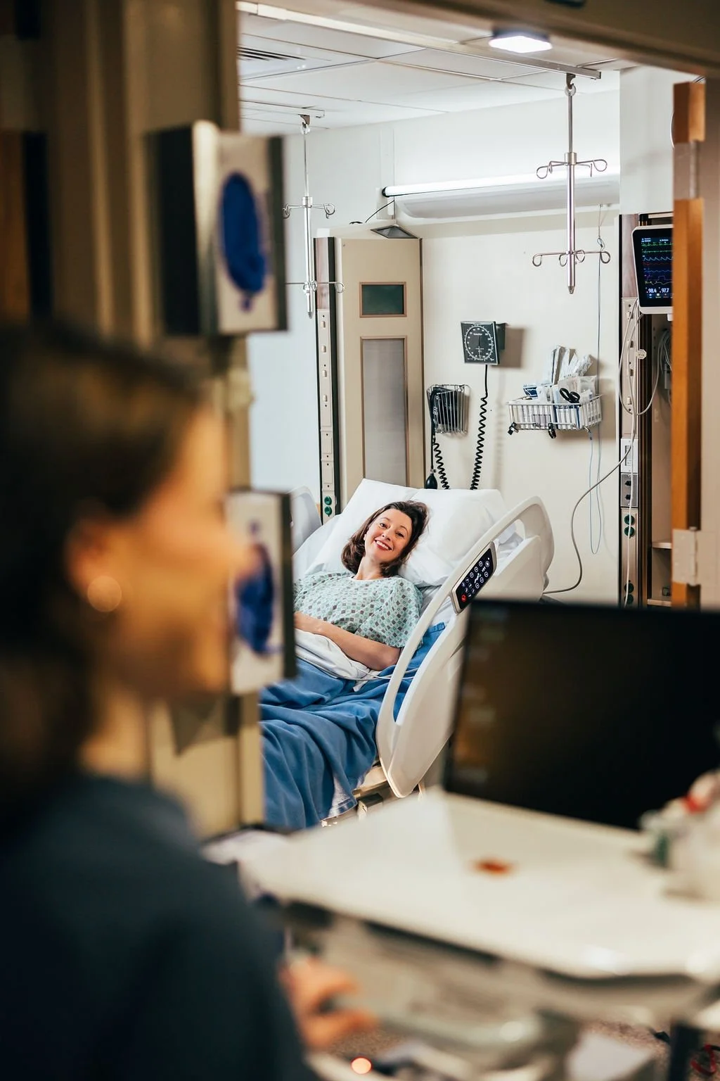 A woman lying in a hospital bed, smiling and looking at a person who is partially visible in the foreground. Medical equipment and monitors are in the background.