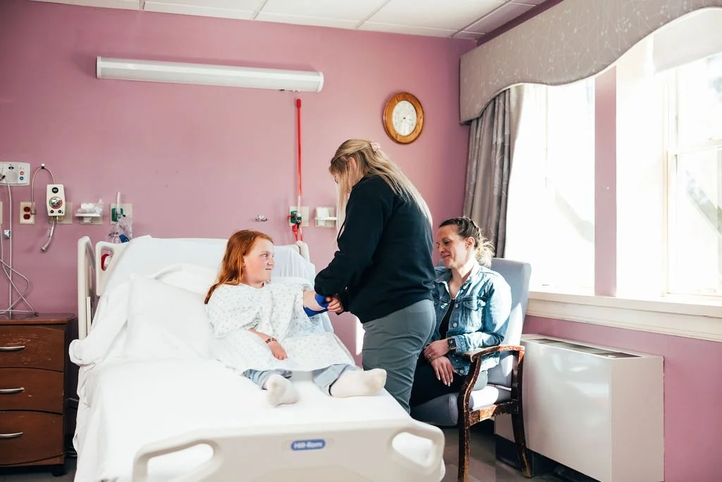 A young girl in a hospital bed talking to a woman standing beside her, with another woman sitting in a chair nearby, in a hospital room with pink walls and large window.