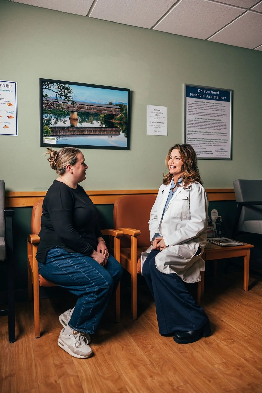 A woman in a black shirt and jeans sitting in a waiting room having a conversation with a healthcare professional in a white coat, who is sitting on a chair. The room has a green wall with a framed picture of a bridge and a river.