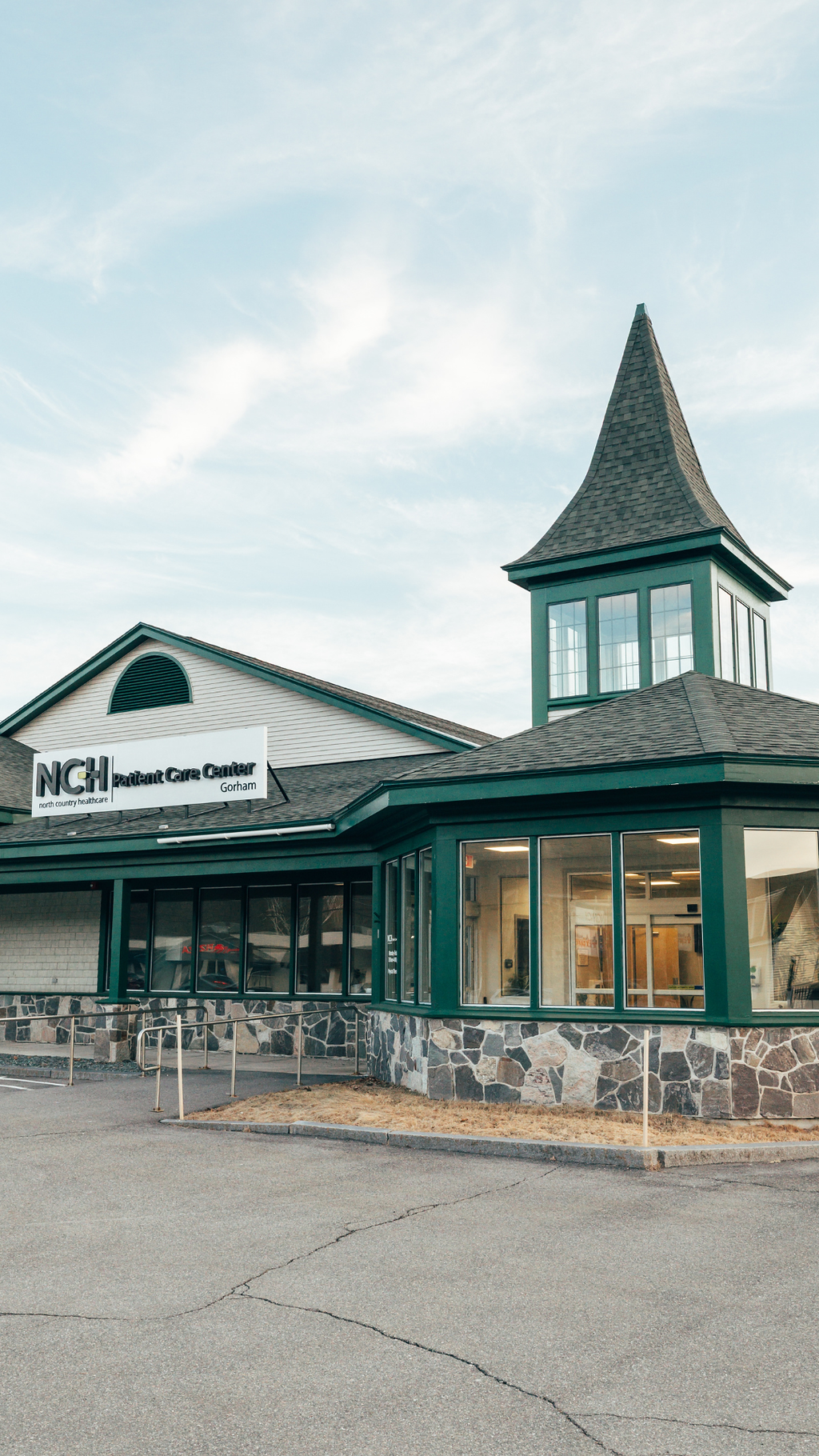 Exterior of North County Healthcare Patient Care Center in Gorham, featuring a green building with a stone foundation and a tall, pointed steeple against a cloudy sky.
