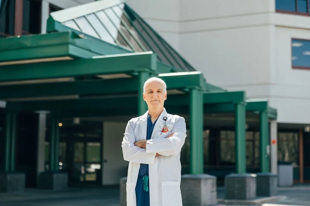 A male doctor in a white coat with a stethoscope standing with arms crossed outside a modern building with green and white exterior.