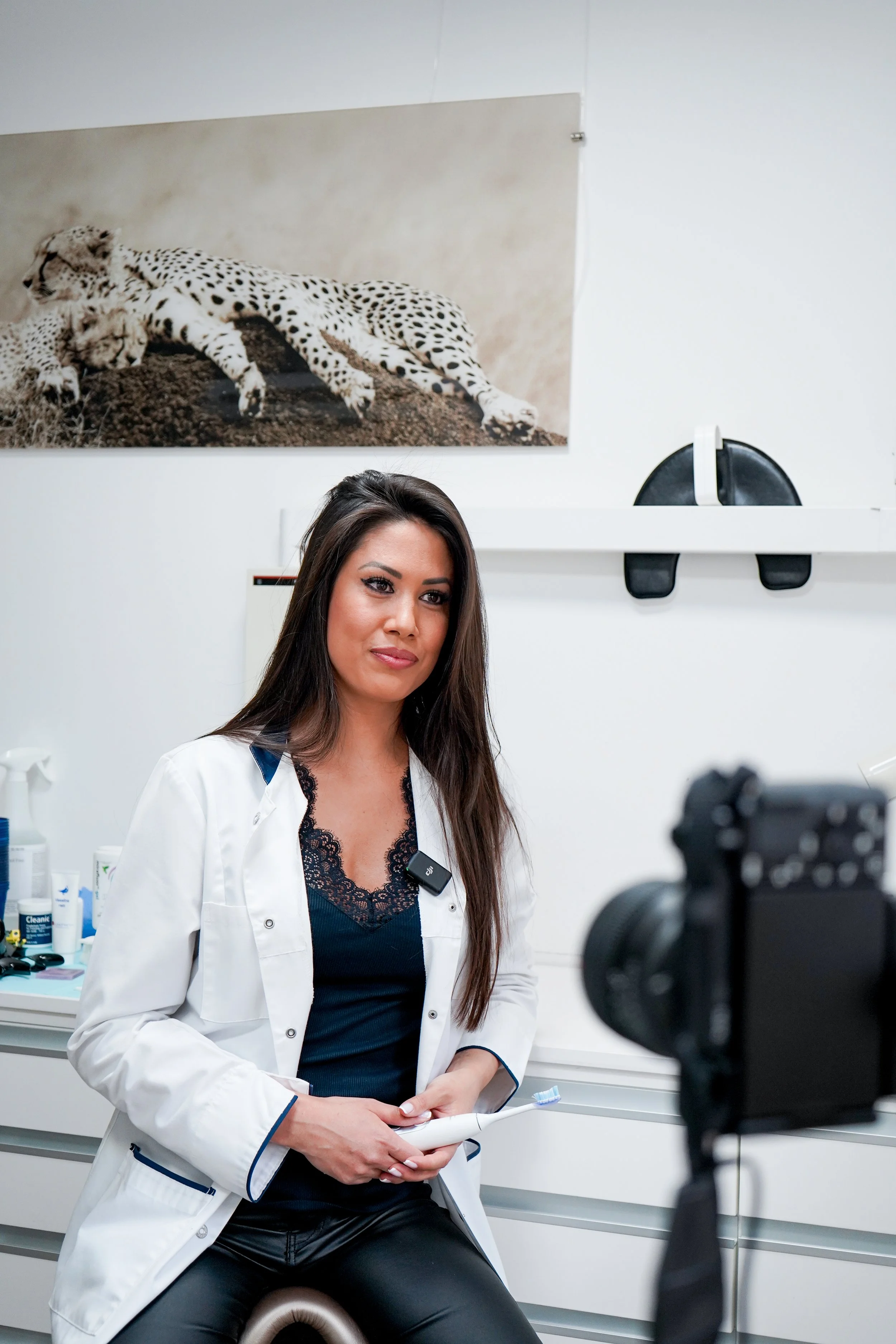 Une femme vêtue d'une blouse blanche, assise dans un environnement médical, probablement un laboratoire ou une clinique, avec un poster d'un léopard en arrière-plan. Elle tient un rouleau de papier ou de documents dans ses mains, et se prépare peut-être à enregistrer des informations ou à filmer une vidéo.