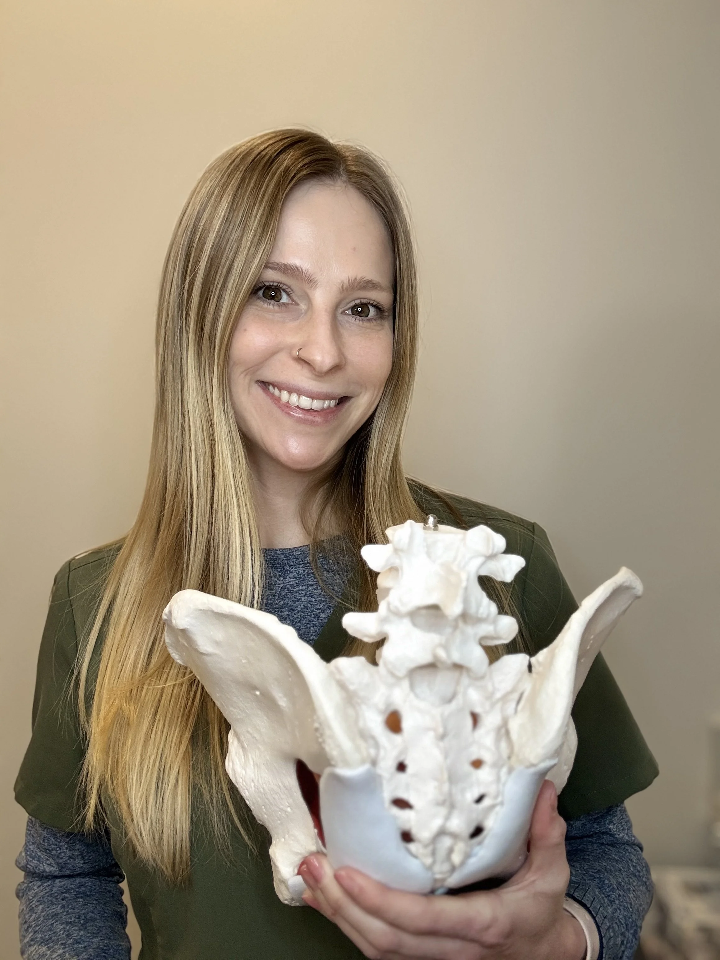A woman with long blonde hair and a nose piercing is smiling at the camera while holding a large anatomical model of a human pelvis.