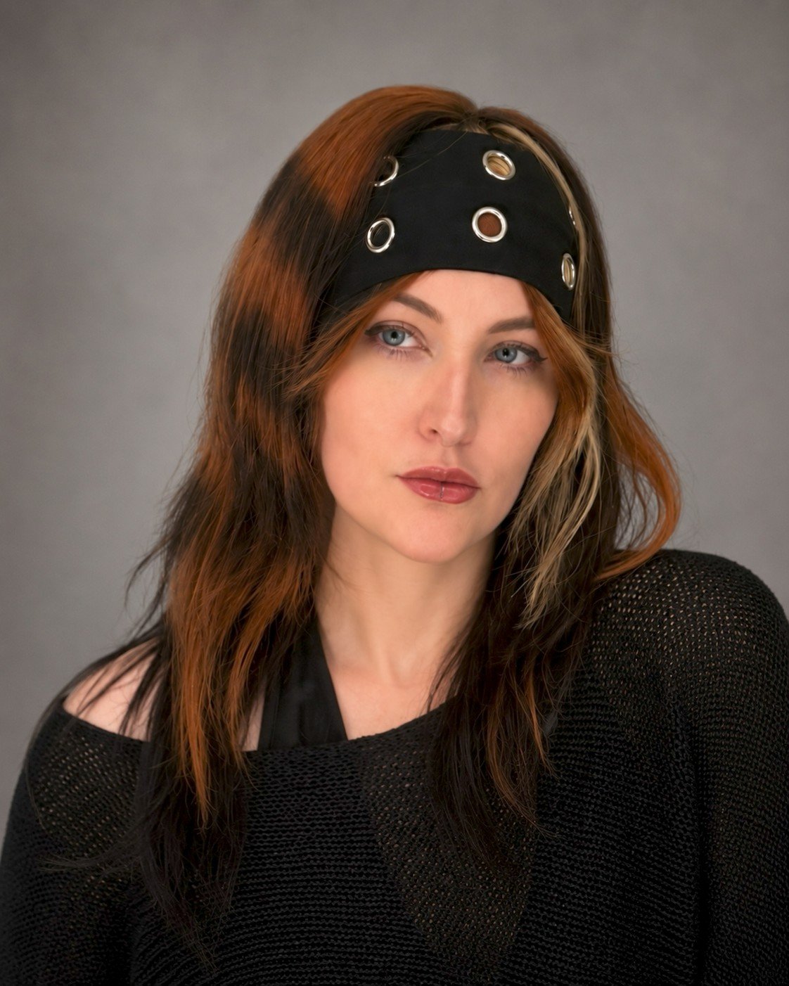 A woman with reddish-brown hair and blue eyes wearing a black headband with metal eyelets and a black knitted top.
