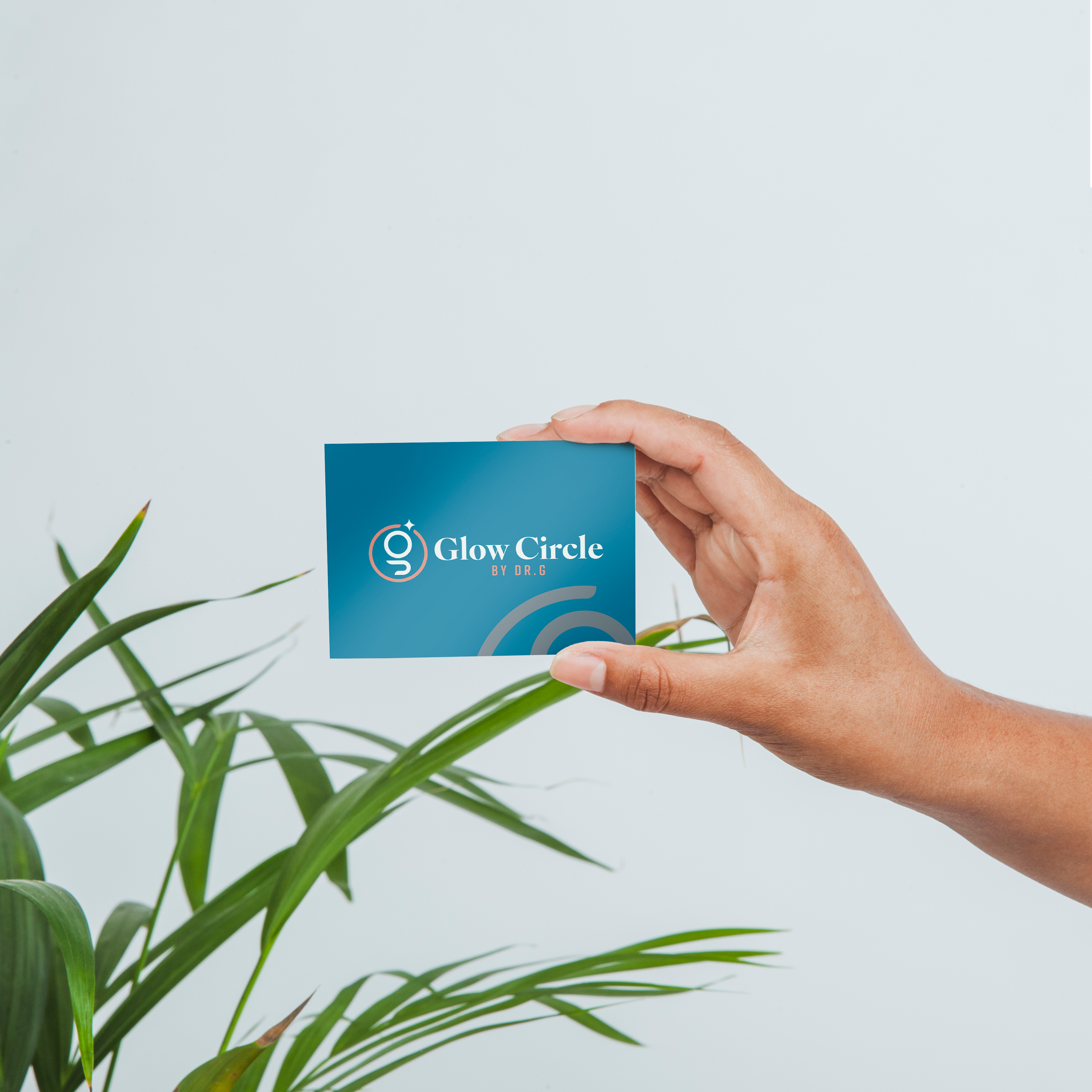 A person holding a blue business card that says 'Glow Circle by Dr. G' with a logo, with green plants in the foreground and a plain white background.