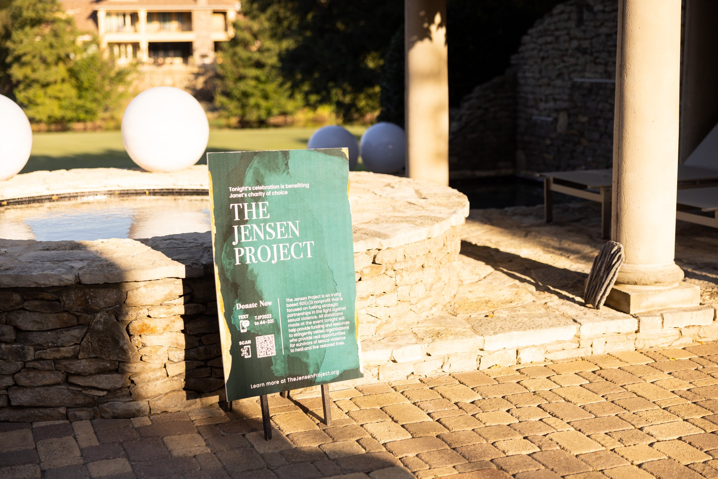 Sign for The Jensen Project charity event outdoors with stone fountain, white spherical lights, and lounge chairs in the background.
