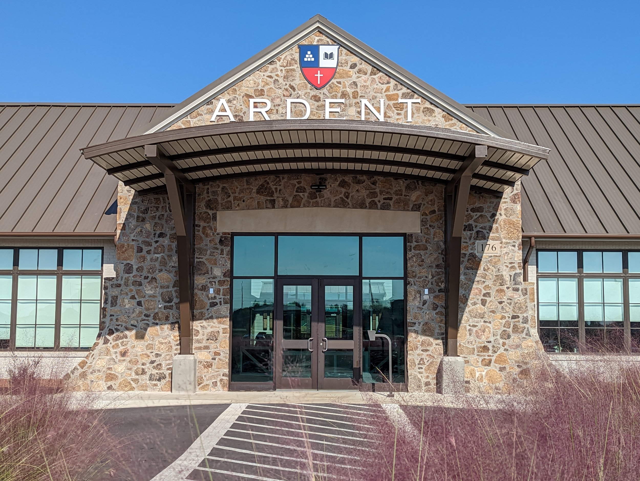 The exterior of a building with a stone facade, glass doors, and large windows, with the word 'ARDENT' above the entrance and a crest featuring books, a cross, and a shield with a cross.