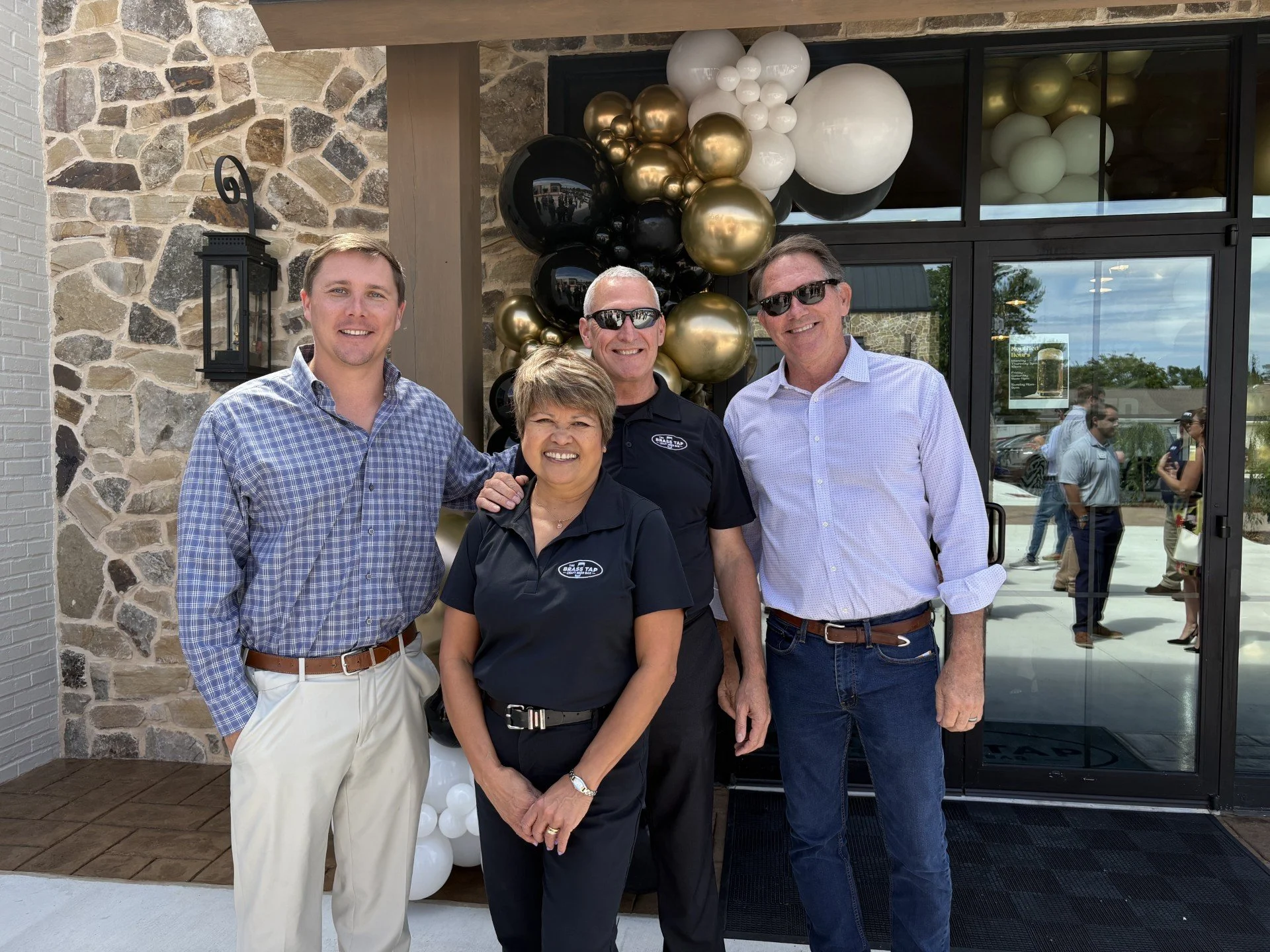 Four smiling people standing outside a building with a stone wall, black and gold balloon decorations, and a glass door.