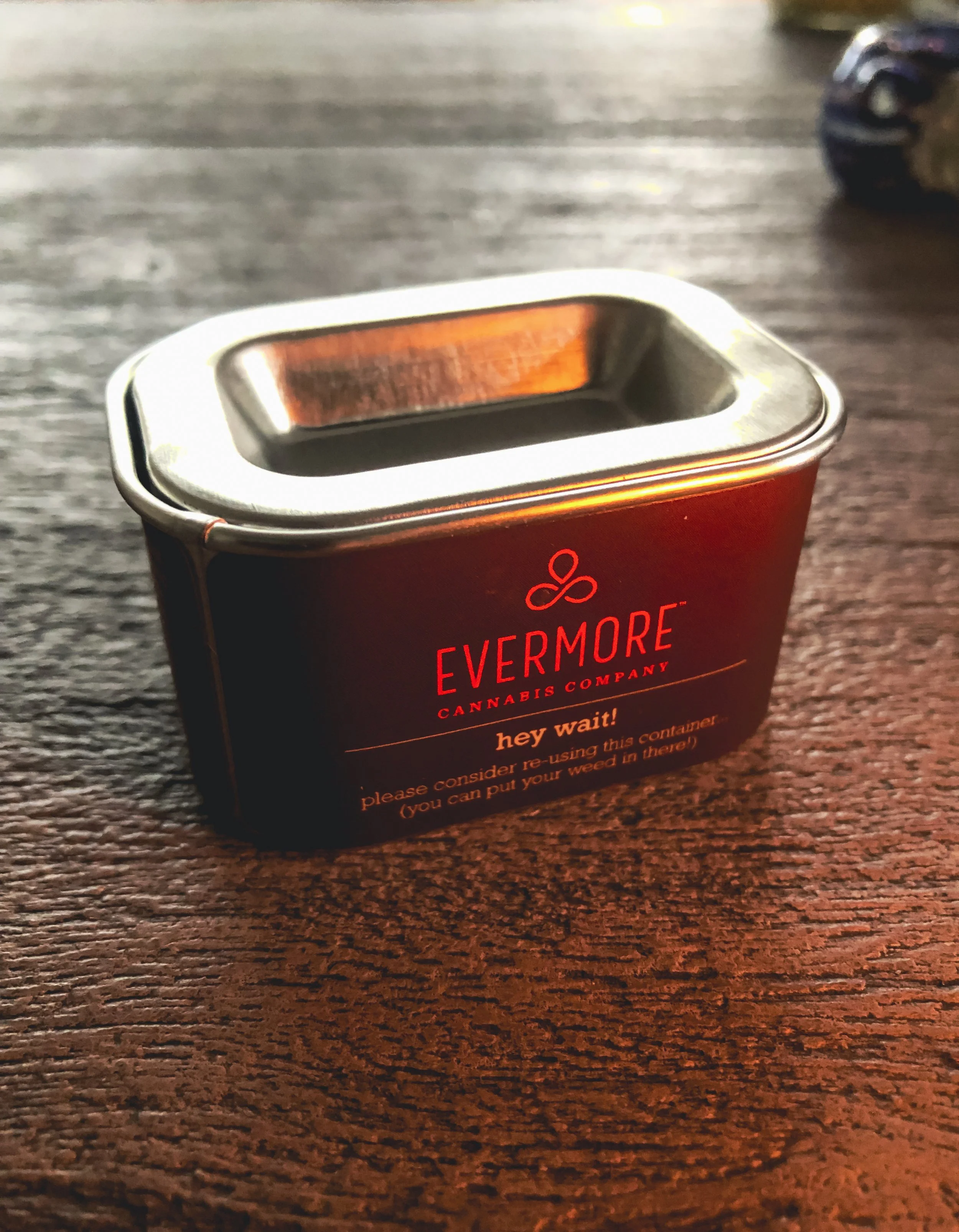 A small black metal container from Evermore Cannabis Company with orange text, placed on a wooden surface, with a small trinket or object partially visible in the background.