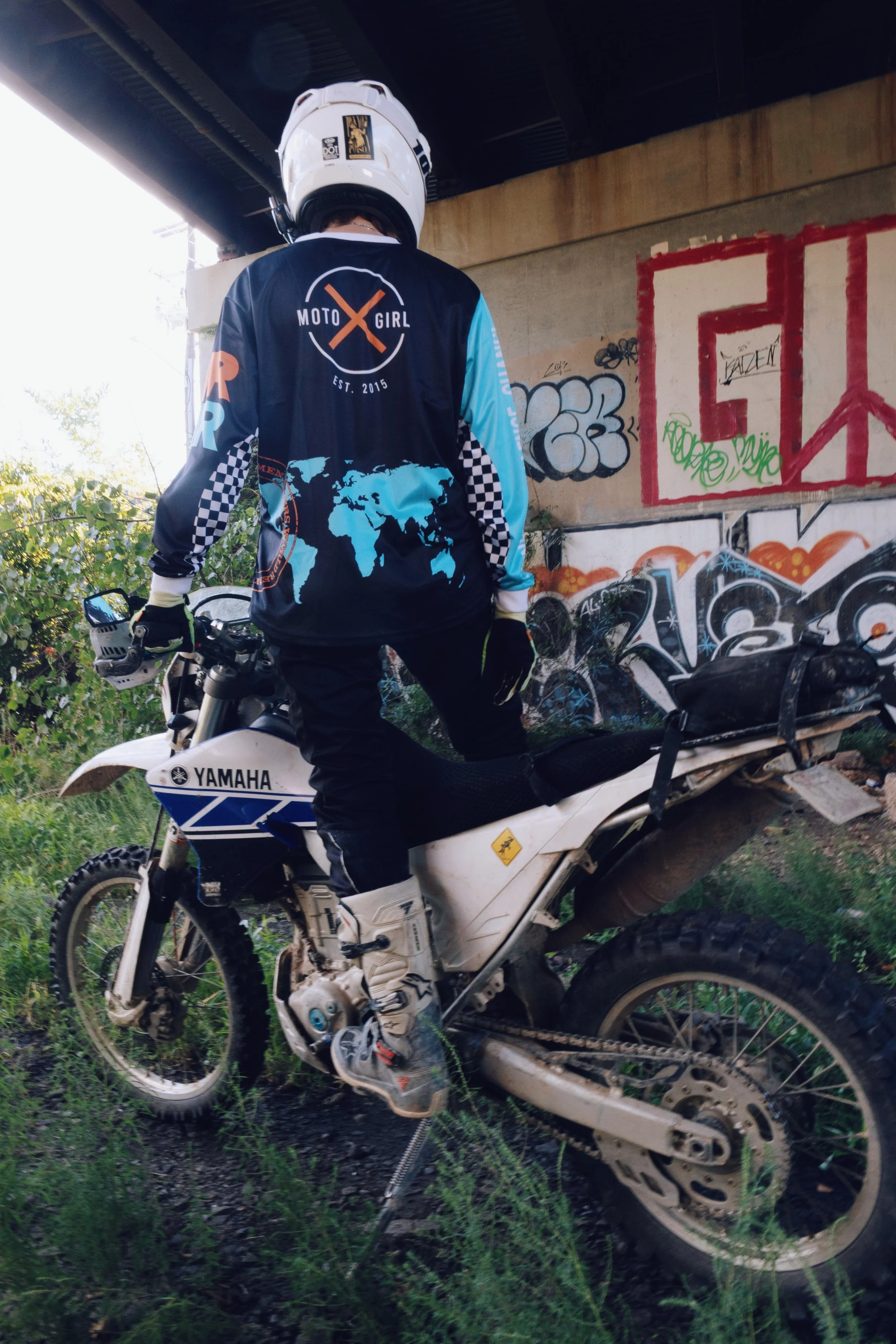 A person wearing a black and blue motocross jersey with 'Moto Girl' and a world map design, sitting on a white Yamaha dirt bike under a graffiti-covered bridge.