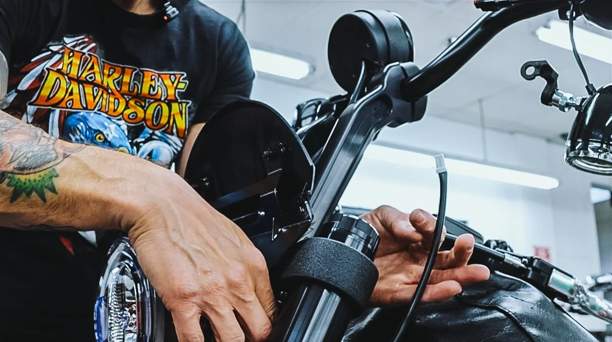 A person working on a motorcycle, with the NAMZ custom wiring extension wearing a black Harley Davidson T-shirt, with tattooed arm visible, in a workshop or garage.