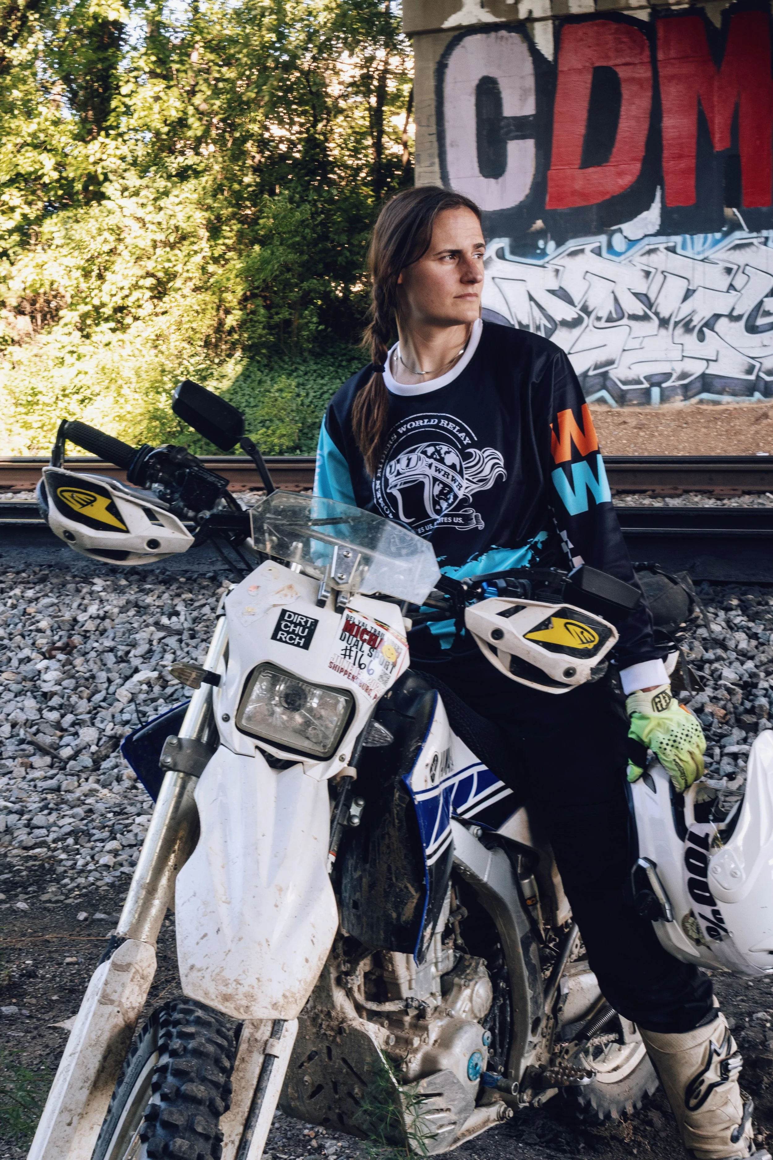 A woman in motocross gear sitting on a dirt bike beside train tracks with graffiti on a wall behind her and green trees in the background.