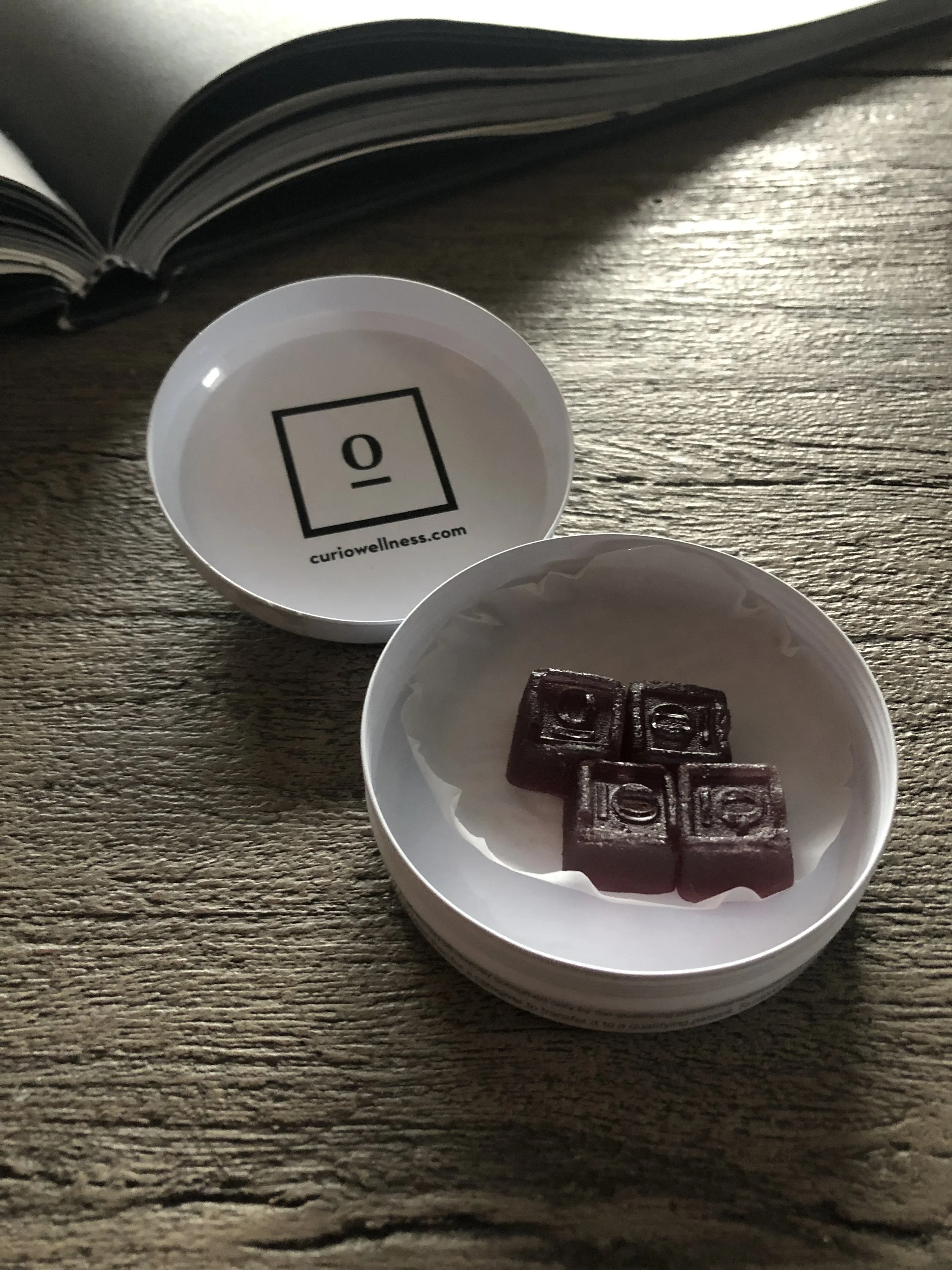Curio Wellness gummies in a white tin on a wood table with a large open book in the background