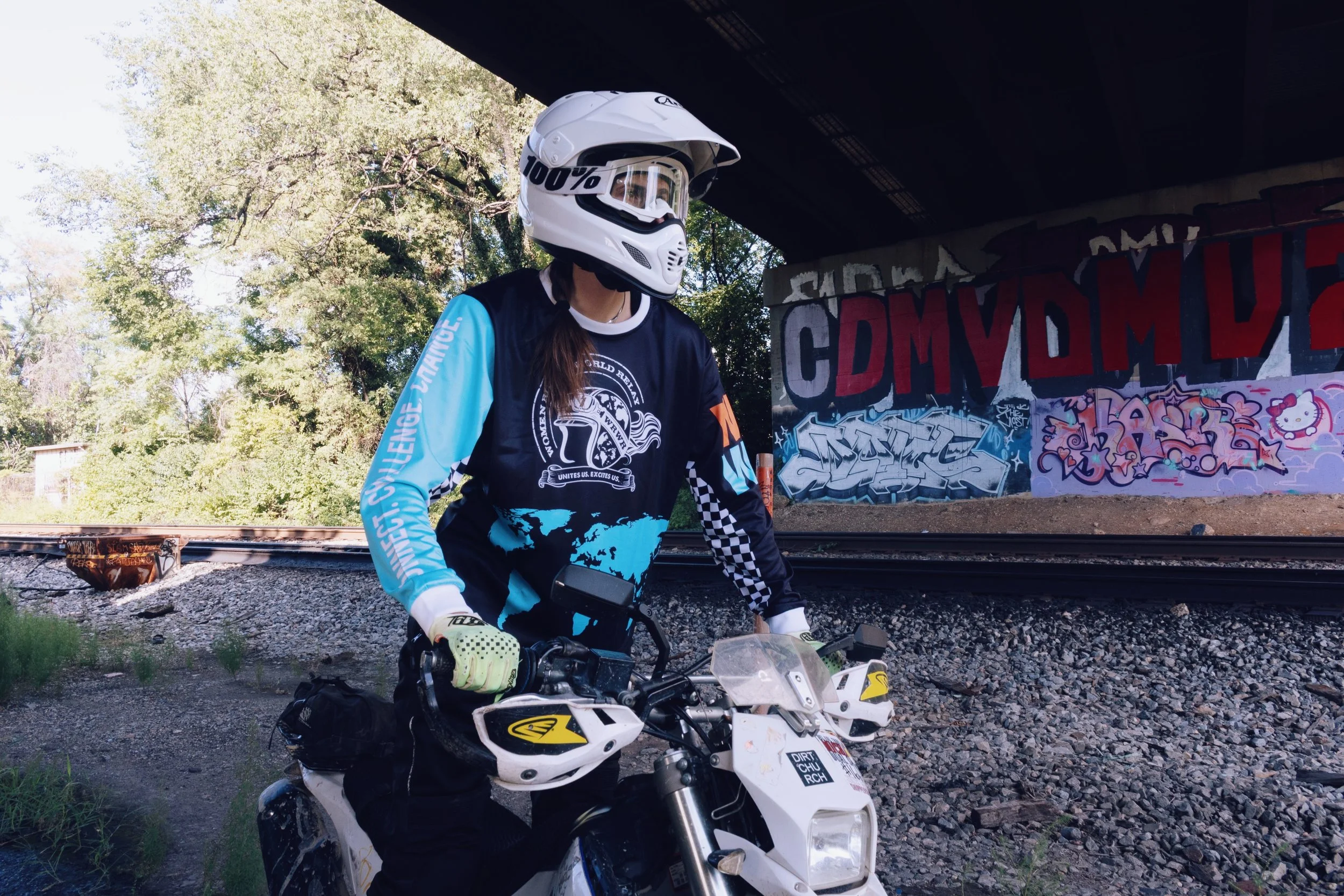 Person wearing a white helmet and black and blue jersey motorcycle riding gear, riding a white dirt bike under a bridge with graffiti, trees, and train tracks in the background.