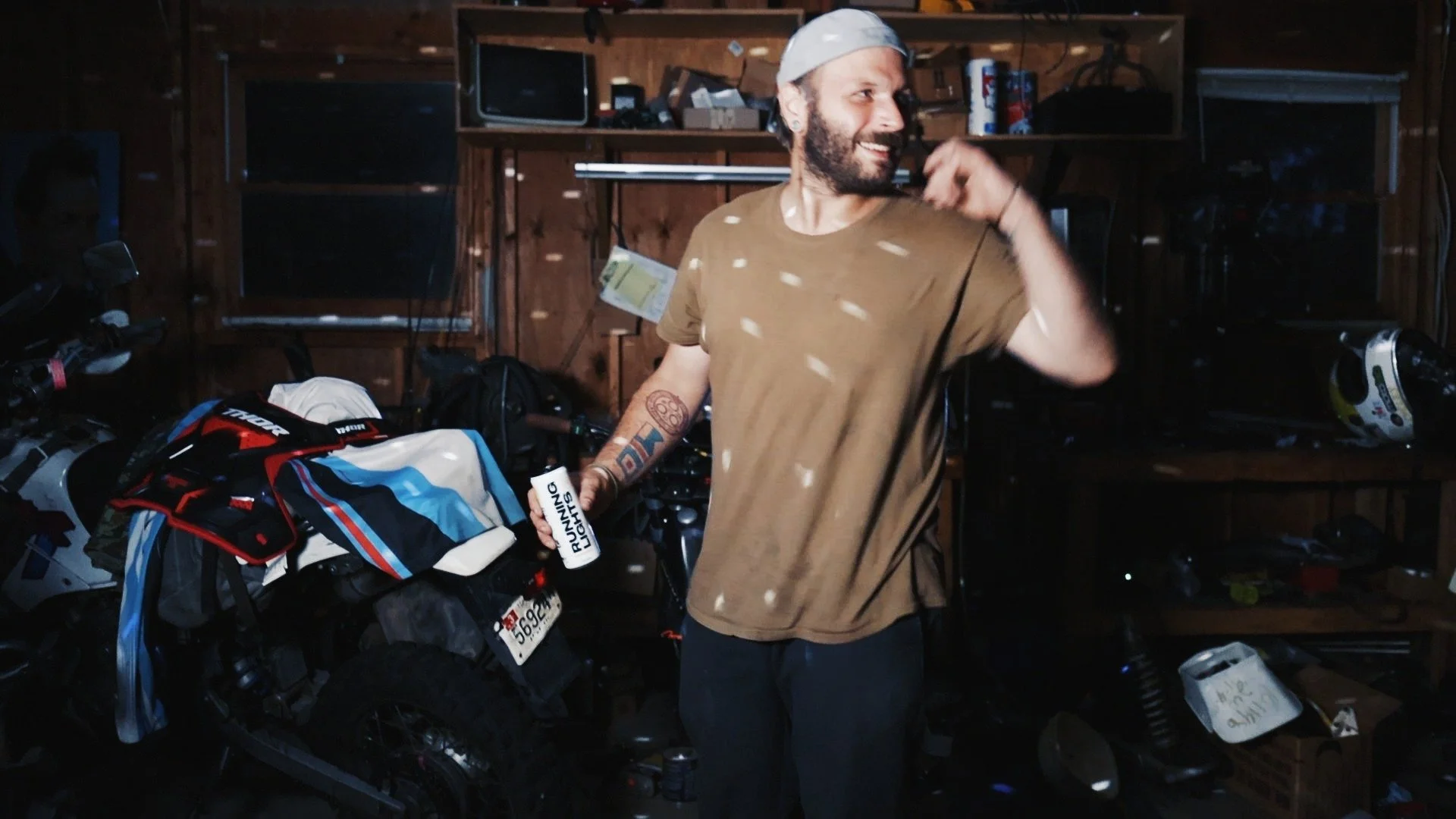 A man with a beard and tattoos on his arm standing in a garage, smiling and holding a can of running light. He is wearing a light-colored cap and a brown t-shirt. The garage has shelves with various items, and a motorcycle with gear is on the left.