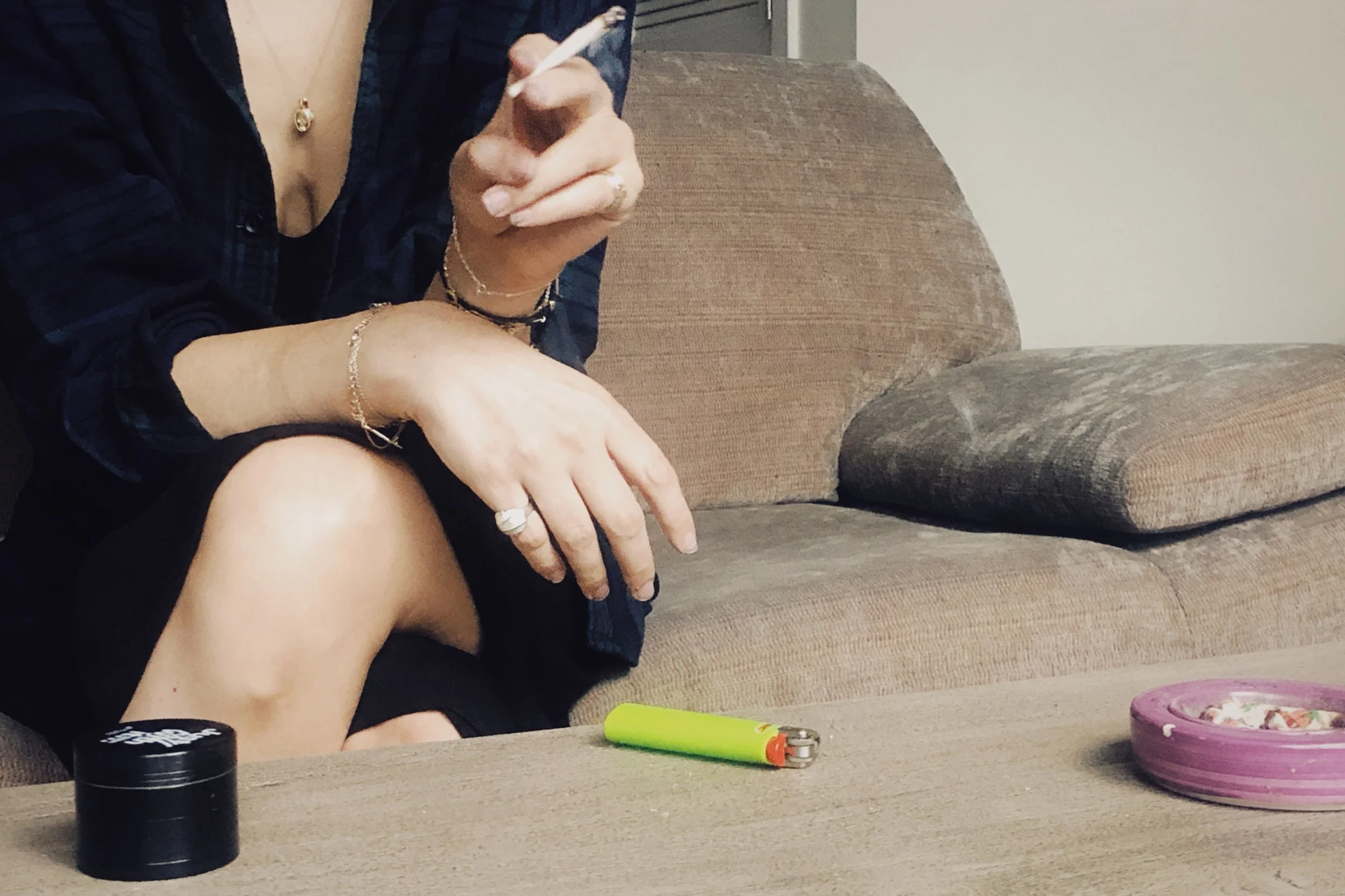 A woman in a black dress holding a pre roll sitting on a beige couch with a black round grinder, light green lighter and purple ash tray in front of her on top of a wooden coffee table.