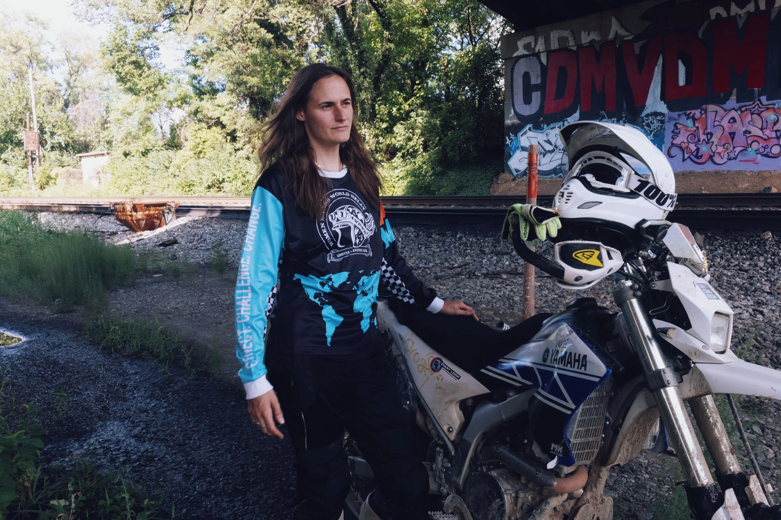 A woman standing next to a white Yamaha dirt bike with a helmet and gloves placed on the bike, outdoors near a railroad track with graffiti on the wall and green trees in the background.