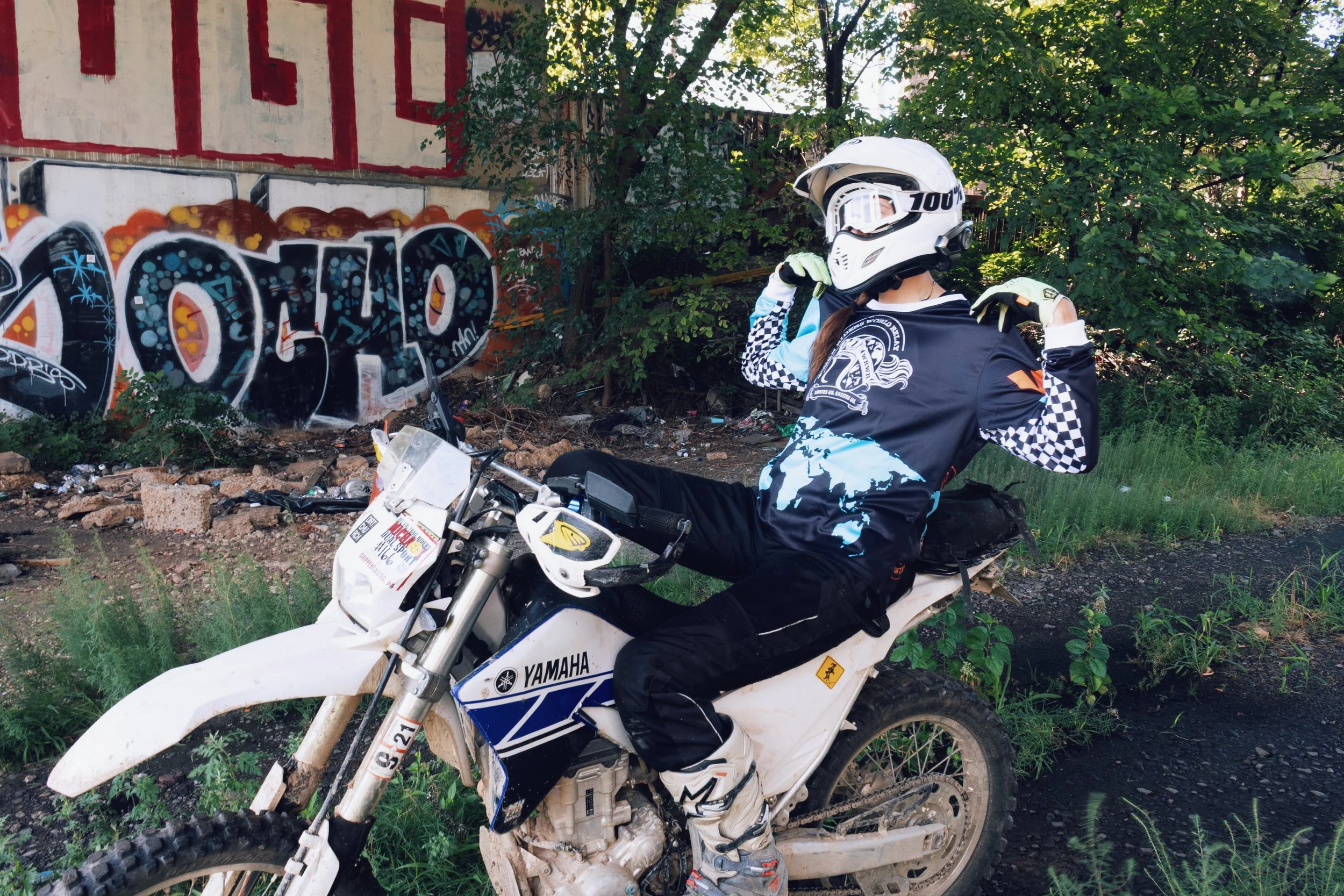 A person wearing a white helmet with goggles, a black and blue motocross jersey, and gloves sits on a white Yamaha dirt bike near a graffiti-covered wall and green trees.