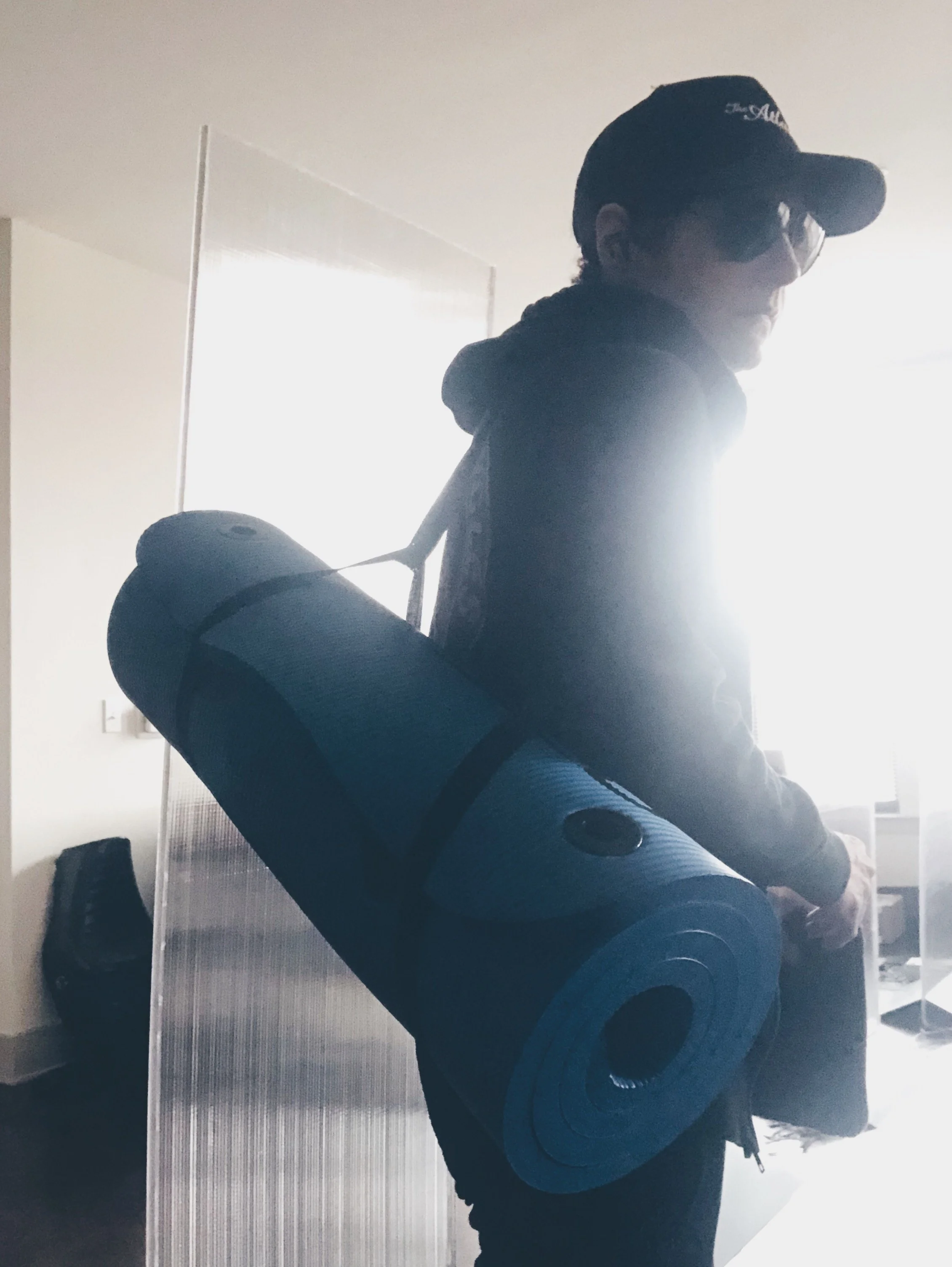 Person carrying a rolled-up blue yoga mat on their shoulder inside a room with sunlight streaming in from a window.