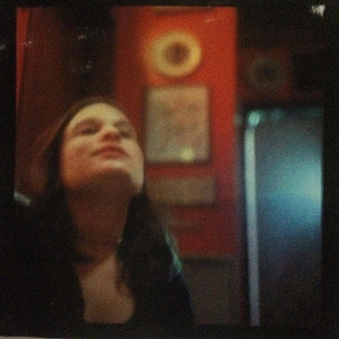 A woman with dark hair looking upward, in a dimly lit room with red walls, a framed record, and a speaker in the background.