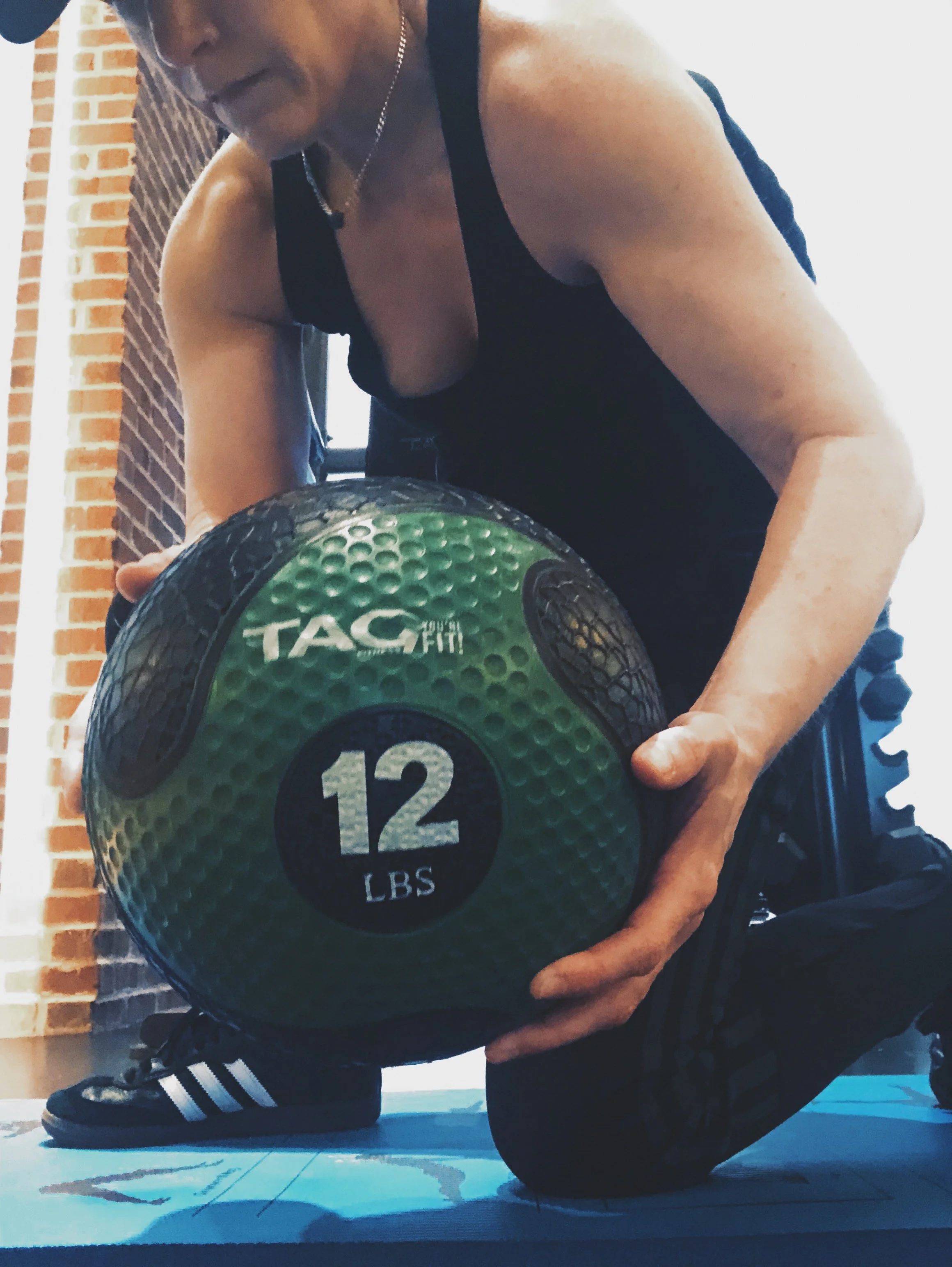 Woman in black workout clothes and baseball hat holding an 12 lb green and black medicine ball from TAG Fitness.