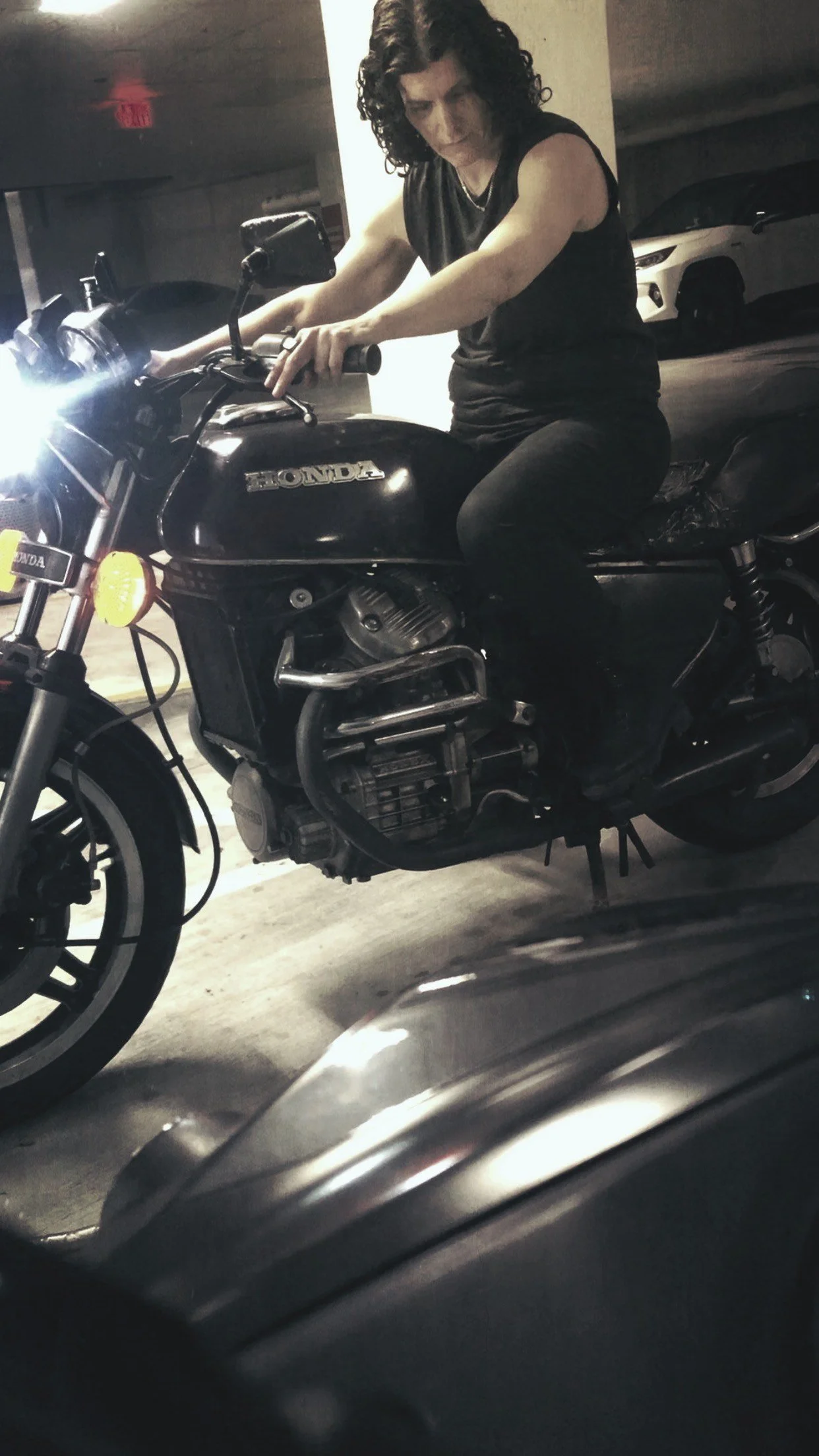 Person with curly dark hair riding a black vintage Honda motorcycle with Rag & Bone muscle shirt, jeans signet ring and combat boots.