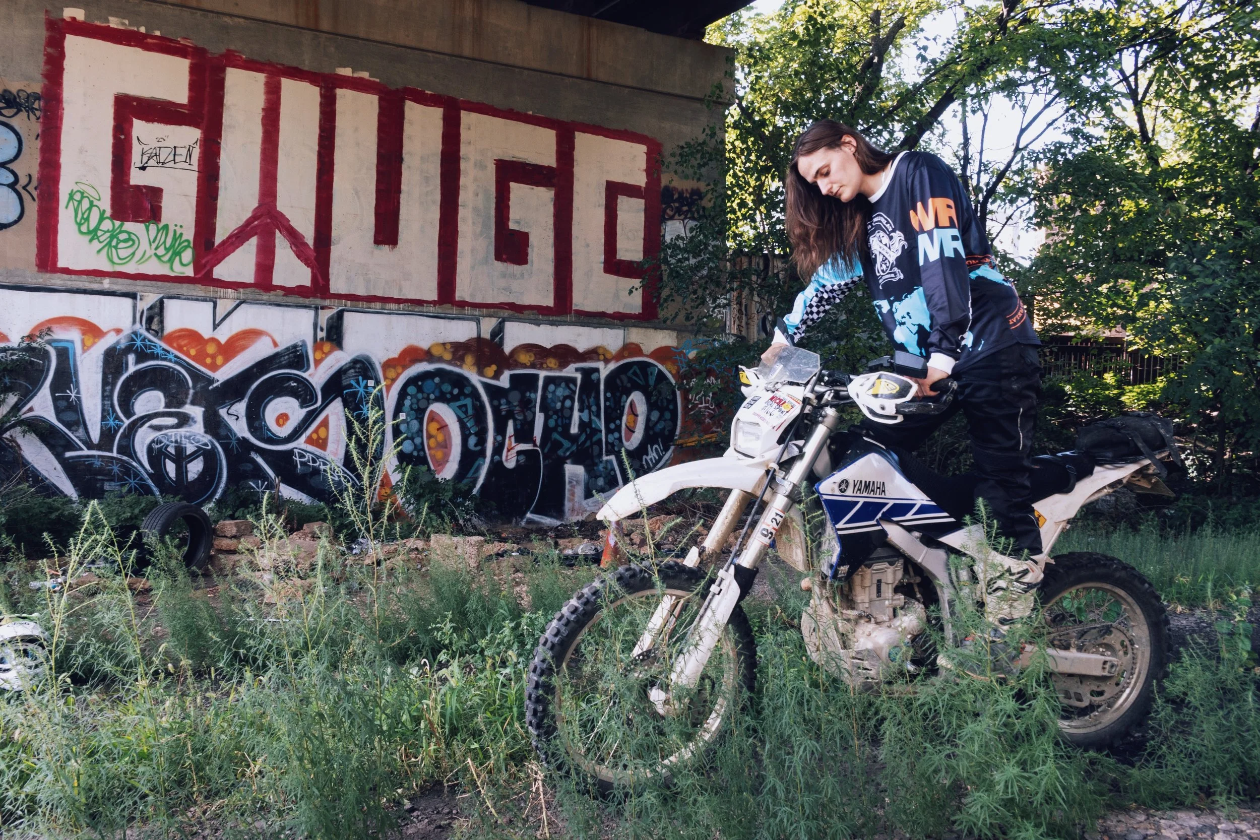 A young woman with long brown hair wearing a black, blue, and orange jacket rides a white Yamaha dirt bike near graffiti-covered walls and trees.