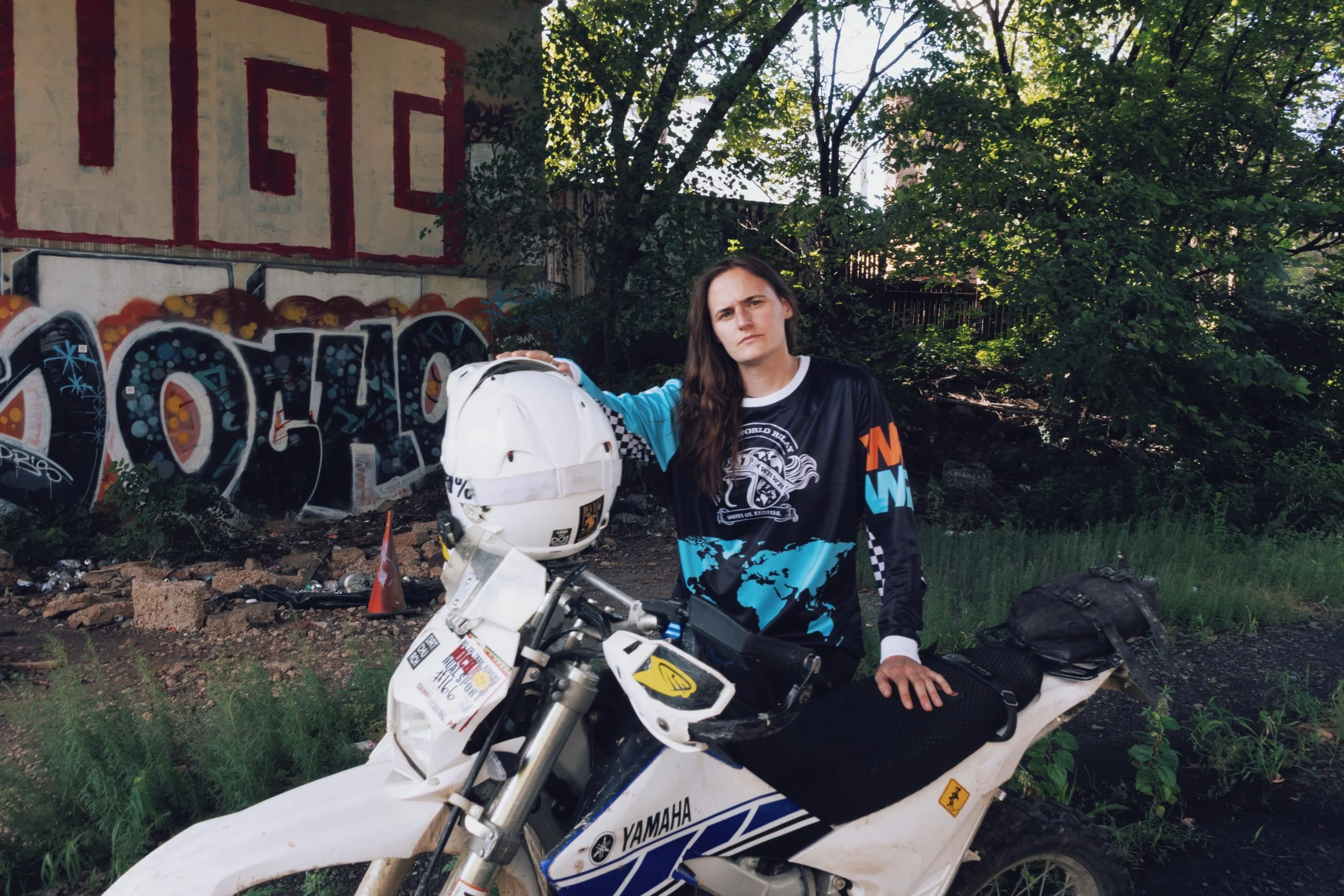 Young woman with long brown hair in a motocross jersey, standing next to a white Yamaha dirt bike in an outdoor area with graffiti-covered wall, trees, and foliage.