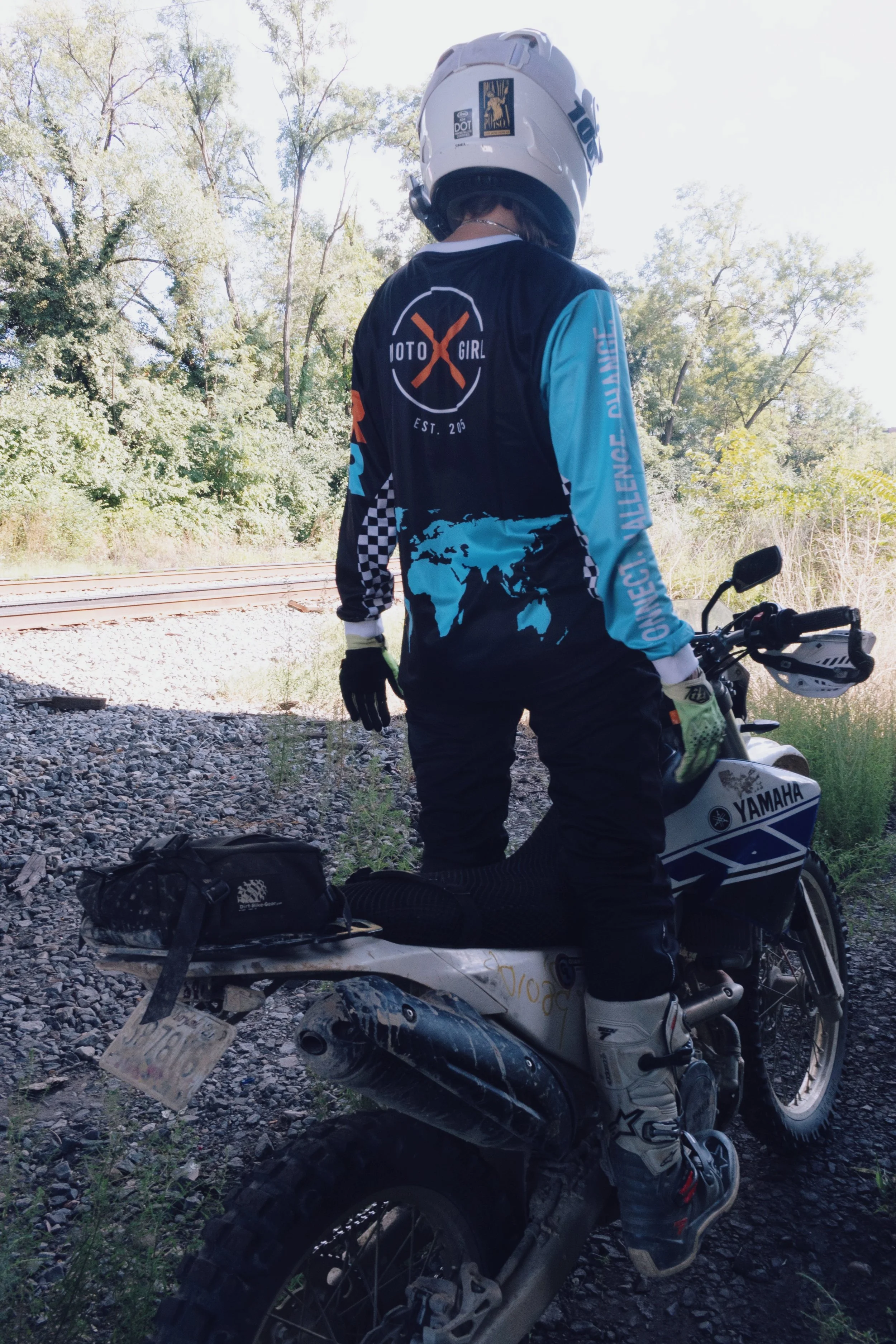 A person standing next to a Yamaha dirt bike on a gravel path, wearing a helmet and motocross gear, with trees and train tracks in the background.